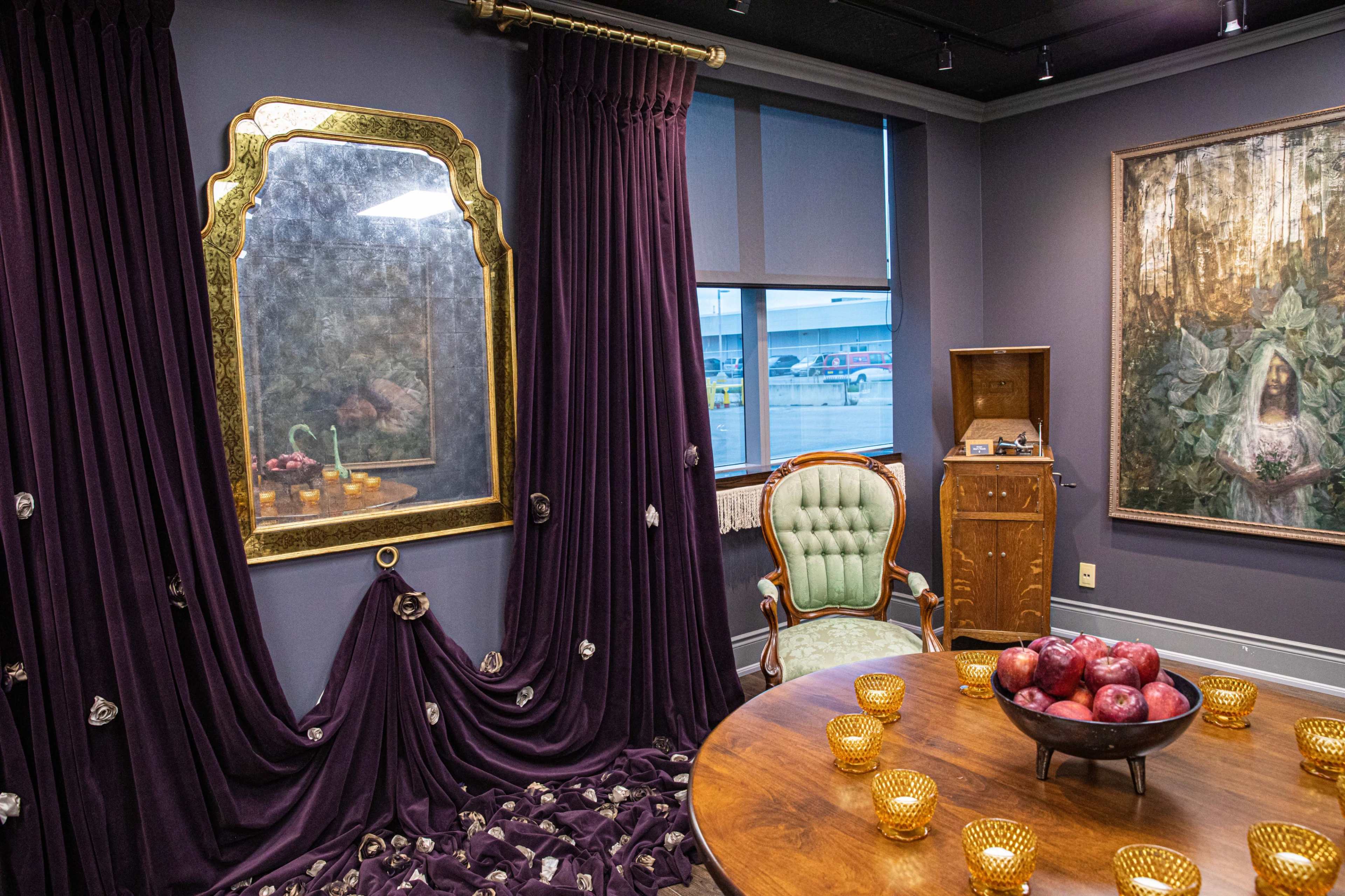 A room features a round wooden table with red apples, a decorative mirror, plush velvet curtains, and an antique chair, all under soft lighting.