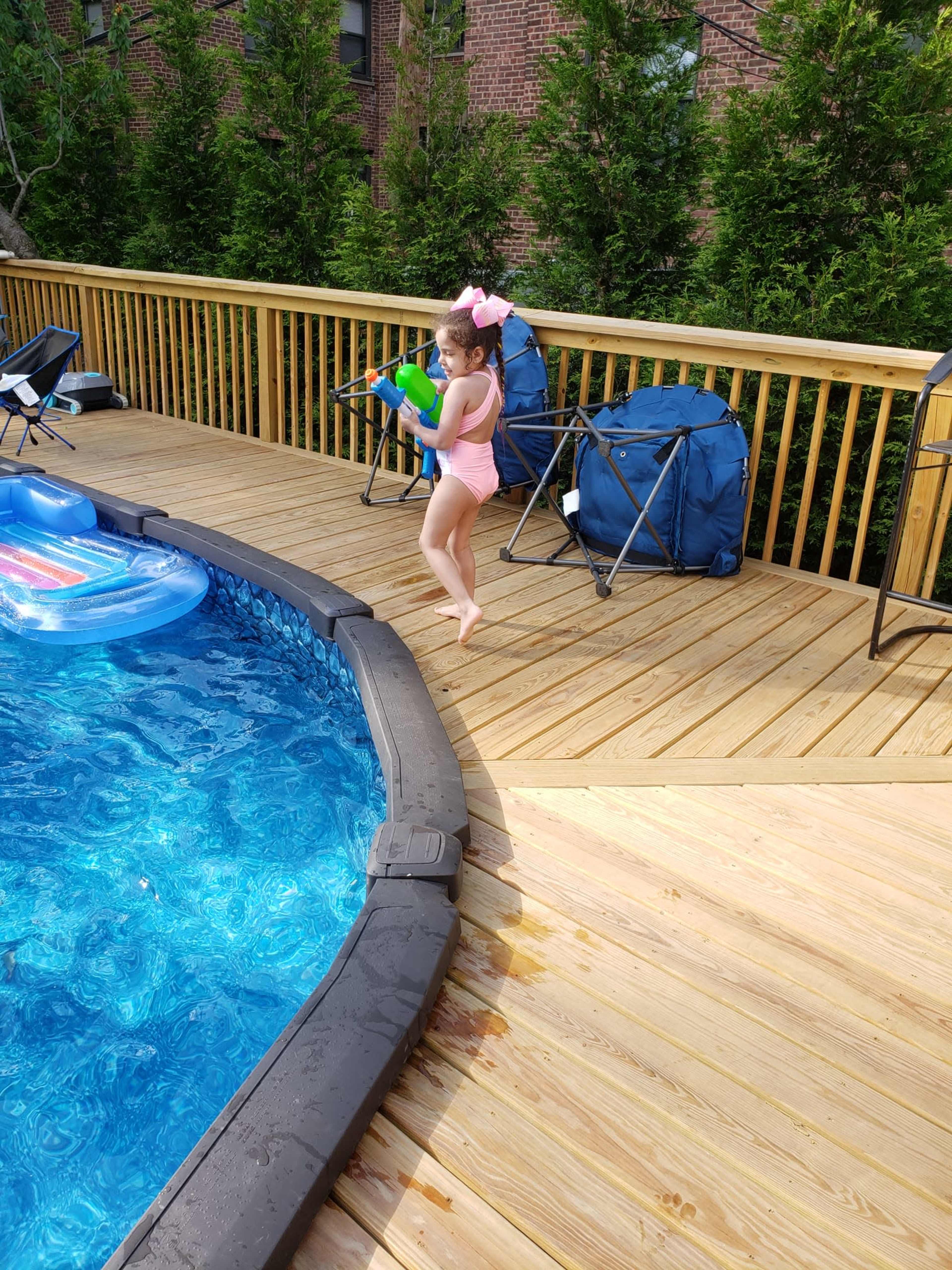 A child wearing a swimsuit and a bow in her hair stands on a wooden deck near a pool, holding a toy.