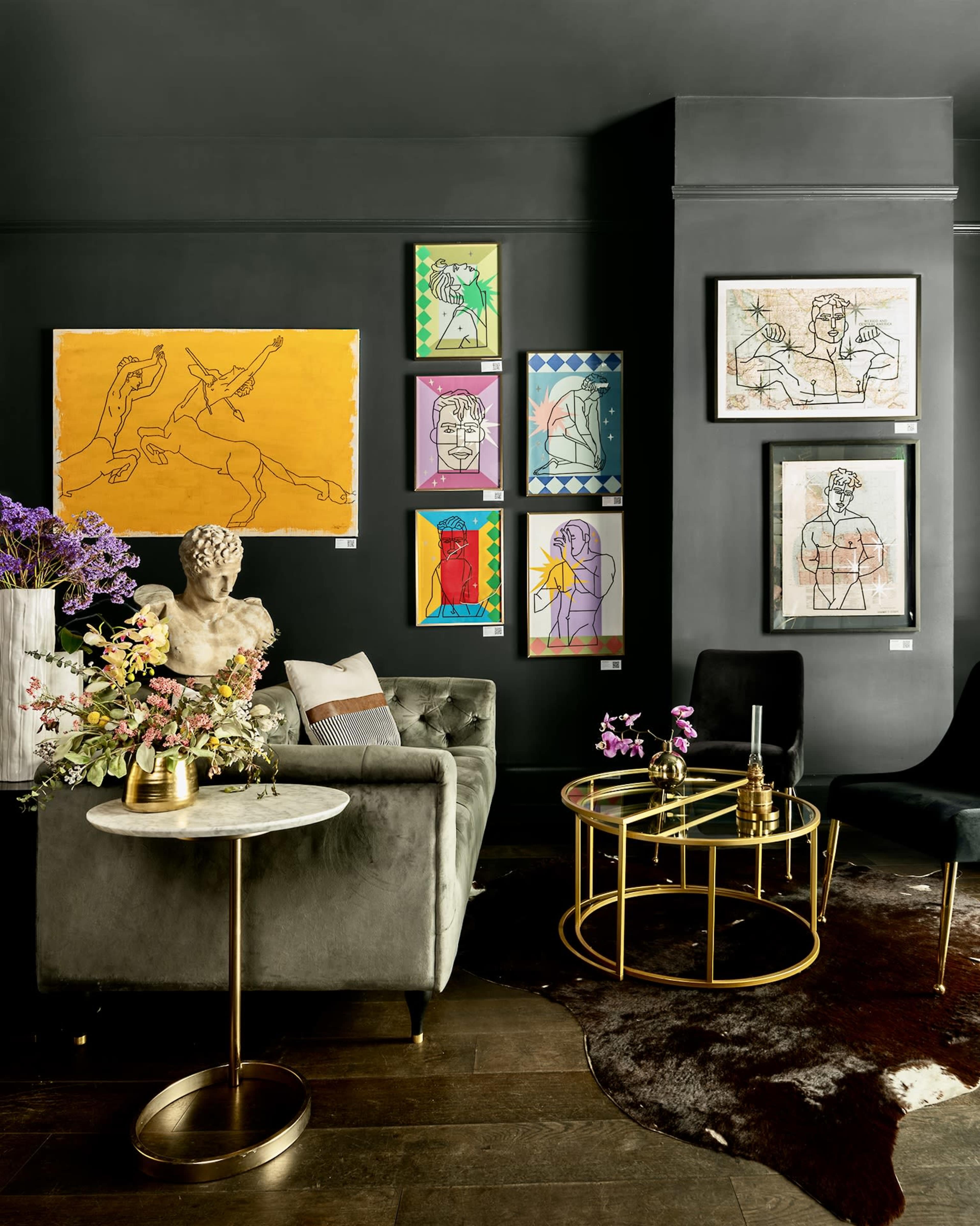 A modern interior features a dark wall adorned with colorful abstract artwork, a marble side table, a plush gray sofa, and a decorative bust amidst floral arrangements.