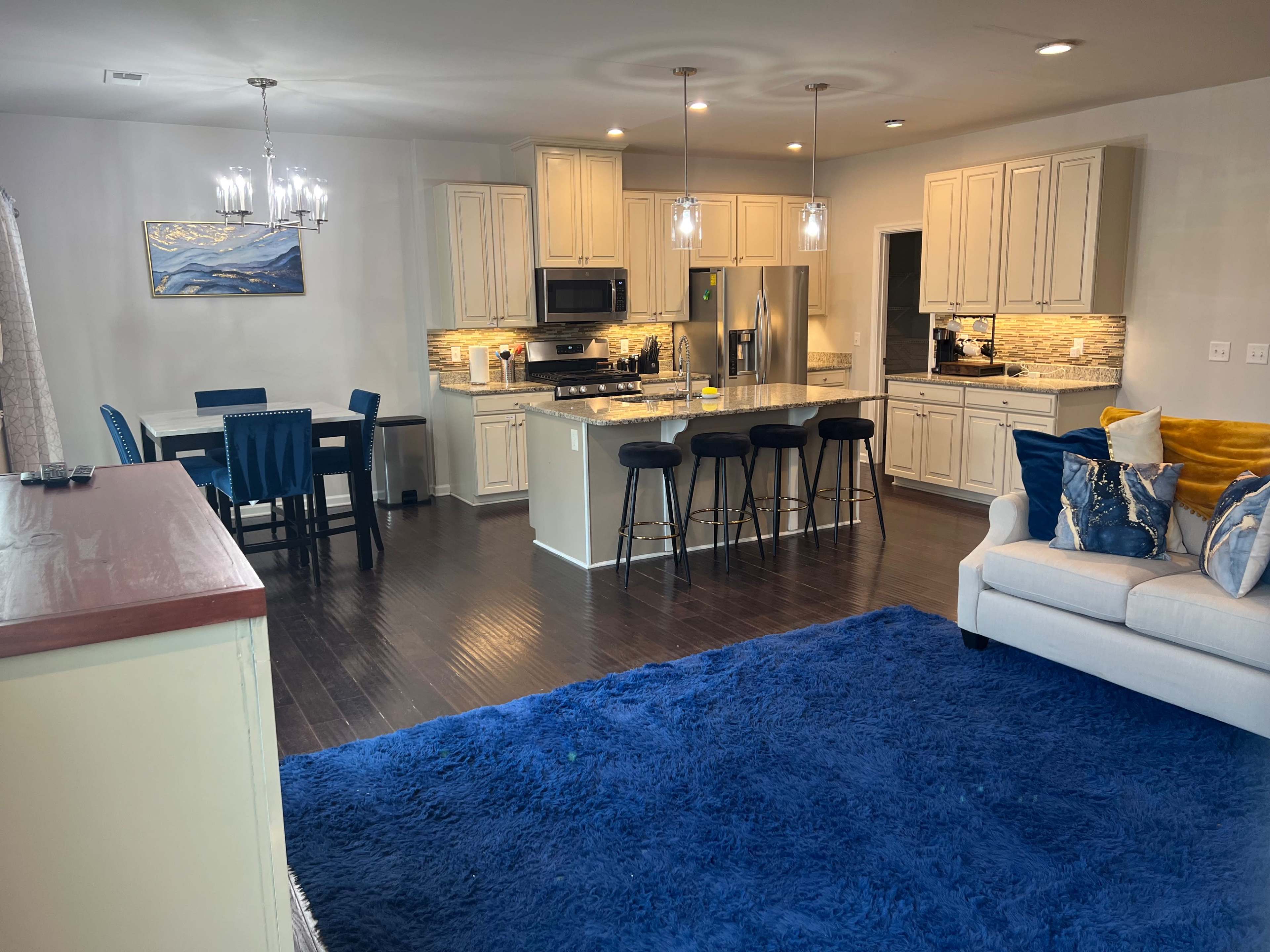 A modern kitchen and dining area with light-colored cabinets, dark flooring, and a blue area rug in the living room.