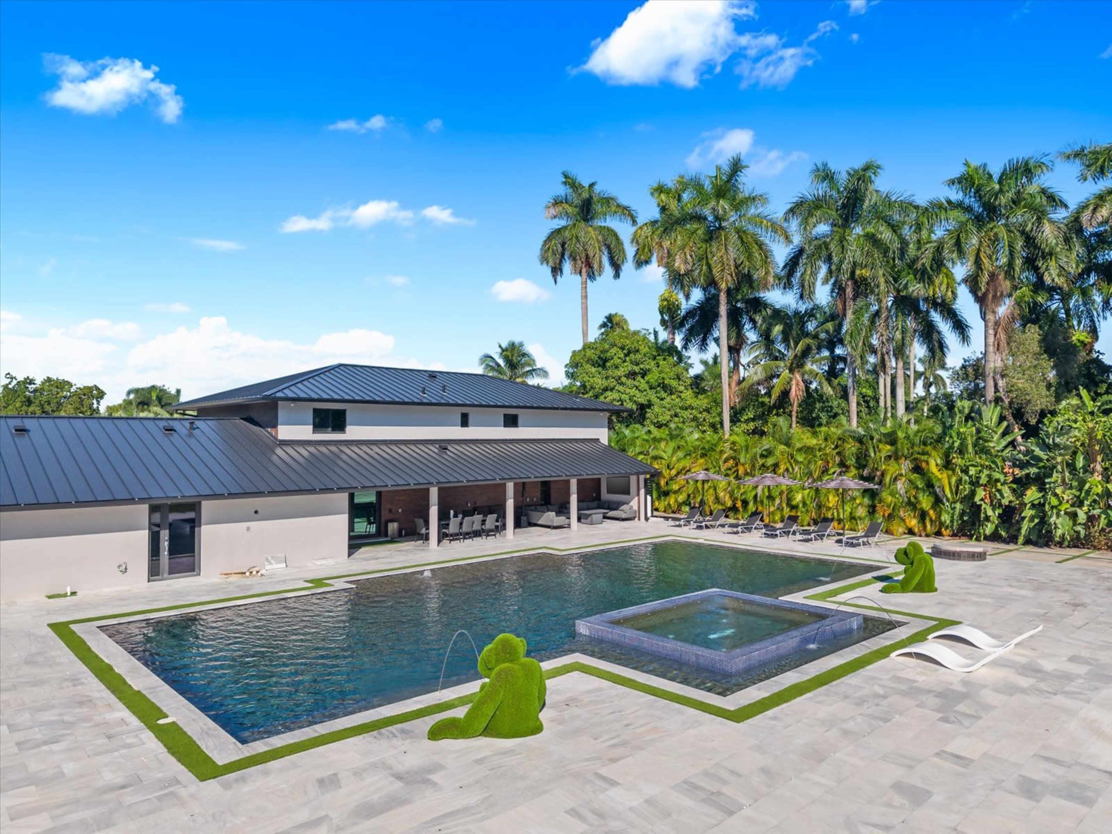 The image features a modern backyard with a large swimming pool, a spa, and palm trees surrounding a contemporary house.