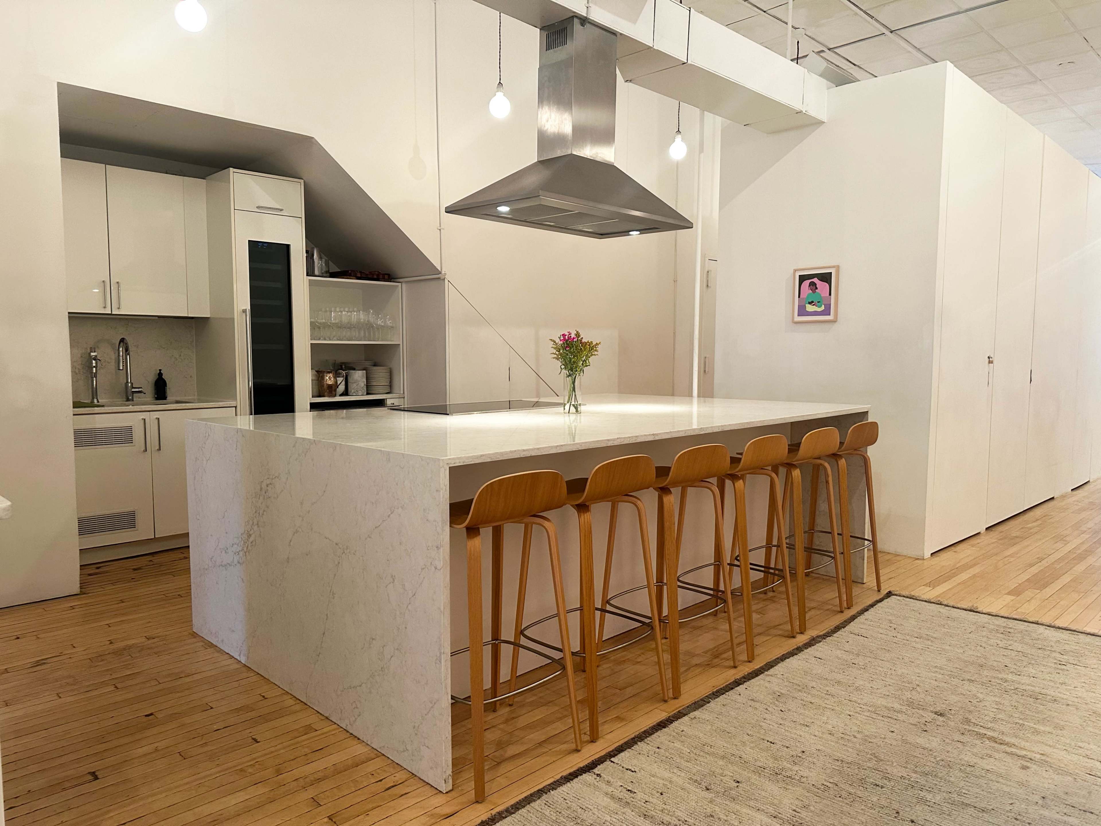 The image shows a modern kitchen with a central island topped with white stone, surrounded by wooden bar stools, and featuring a stainless steel hood above the cooktop.