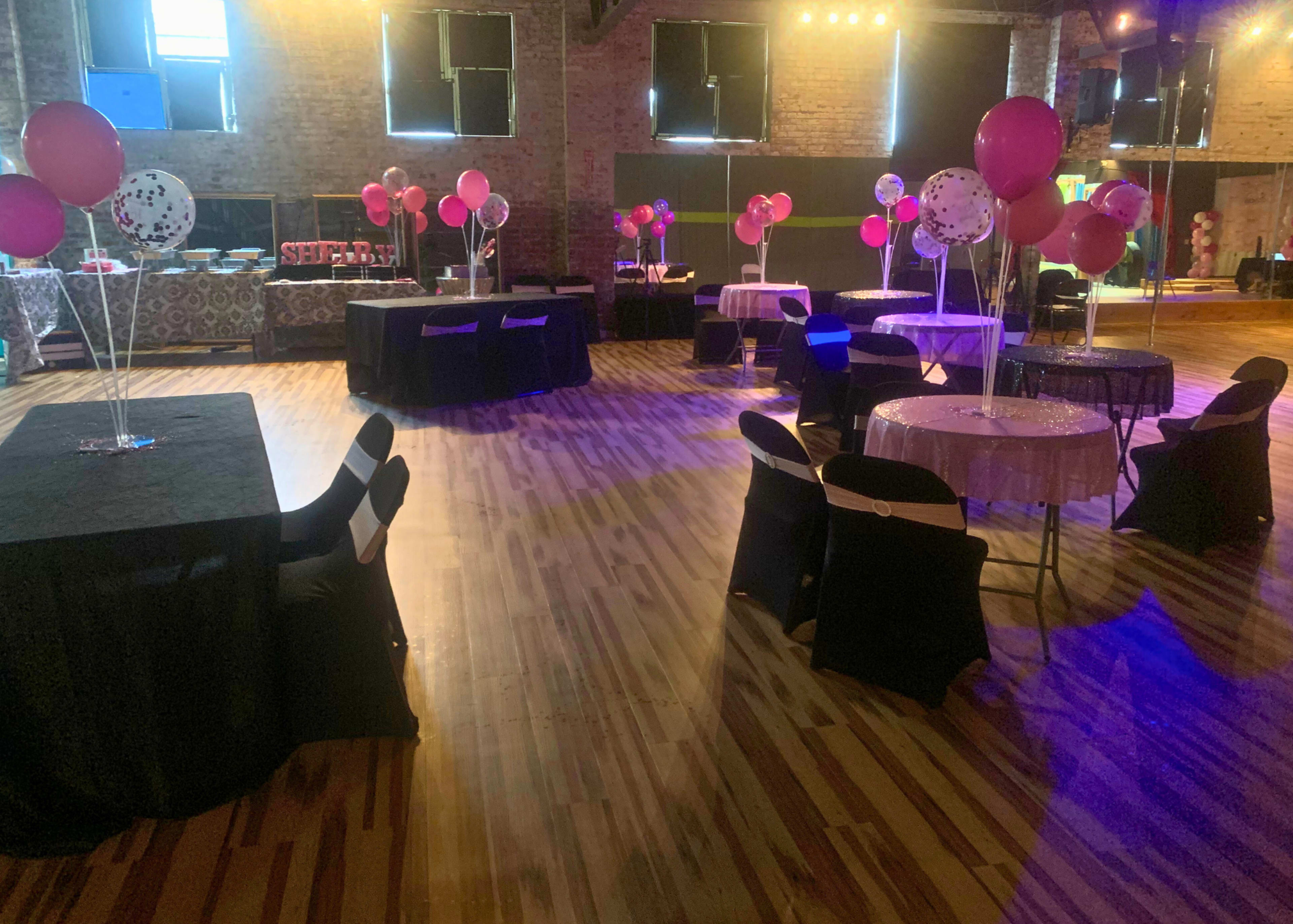 A decorated event space with tables and chairs, featuring balloons and a wooden floor.
