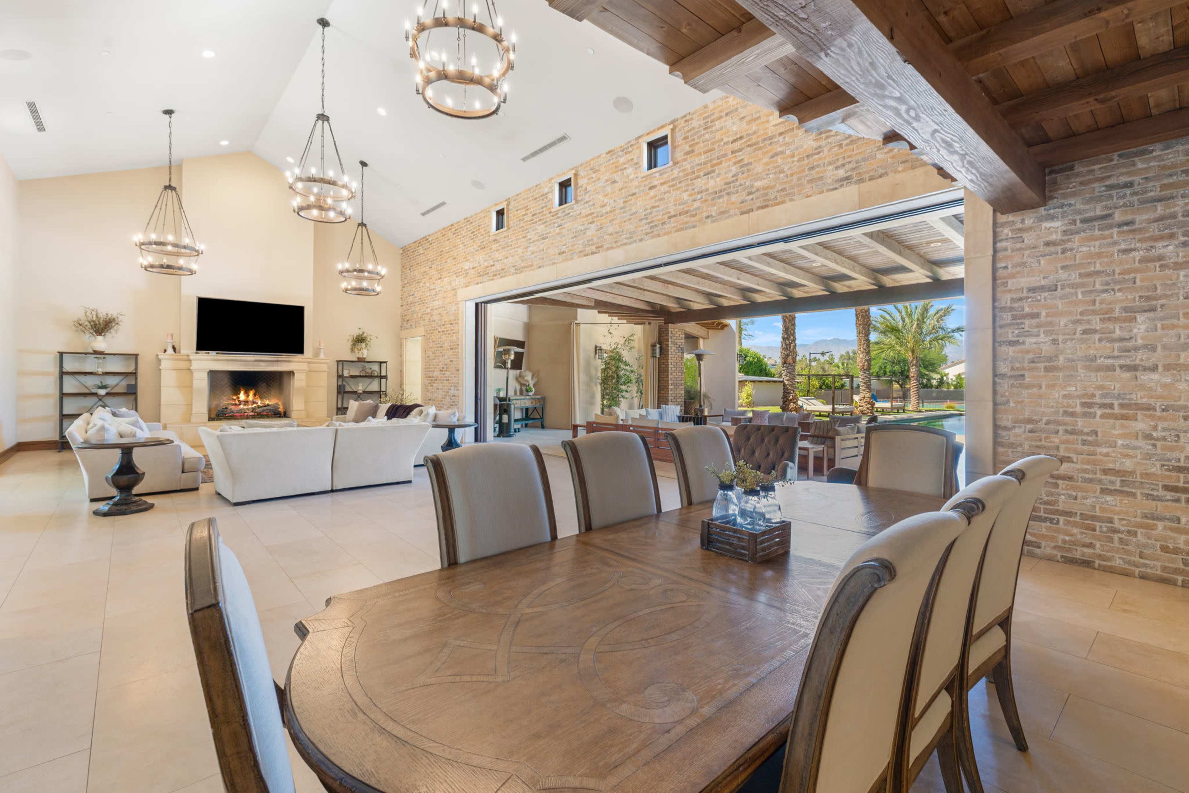 The image shows a modern dining area with a wooden table surrounded by upholstered chairs, open to a spacious living room with a fireplace and large glass doors leading to an outdoor area with palm trees.