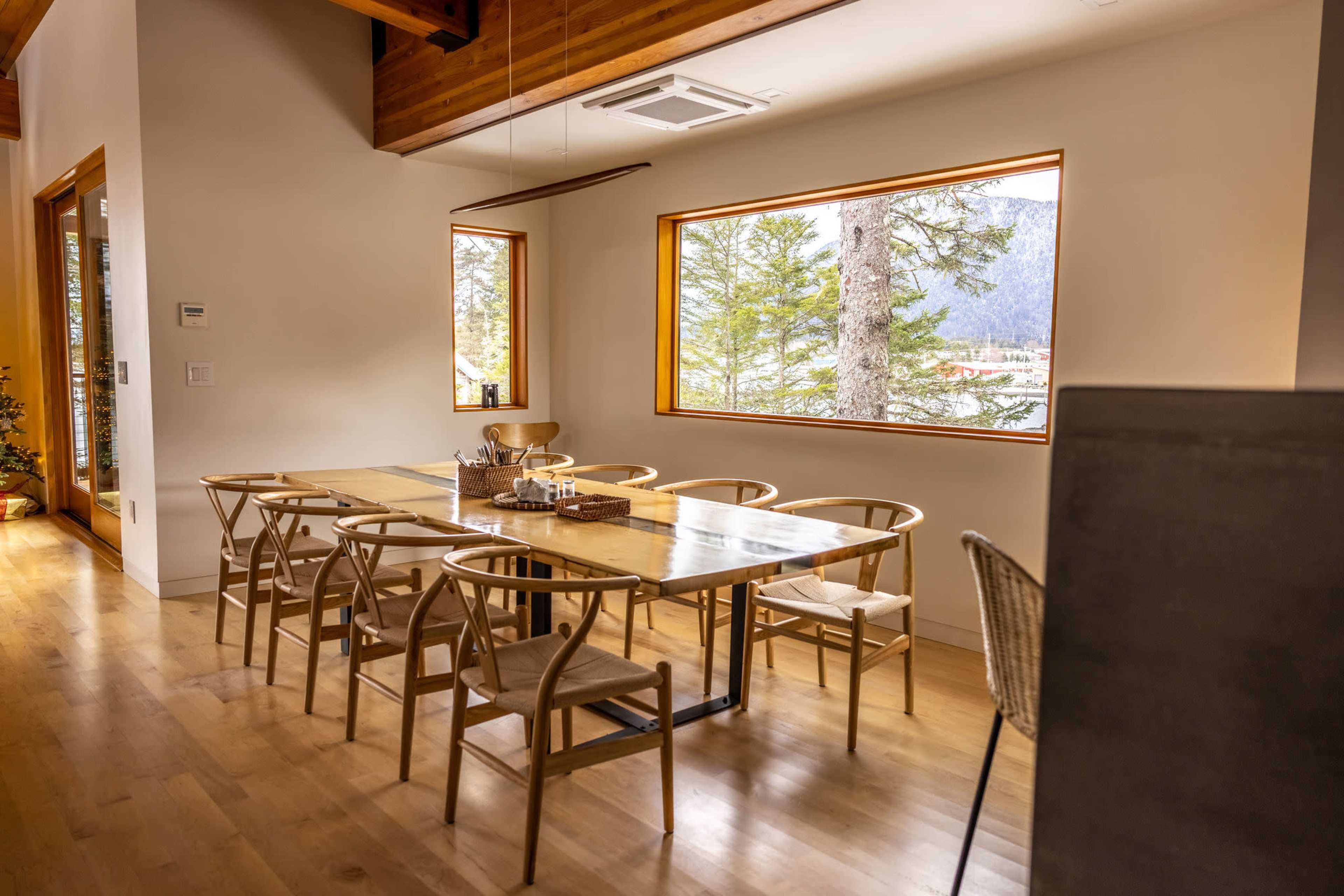 A dining area features a long wooden table surrounded by chairs, with large windows displaying a view of trees and a lake.