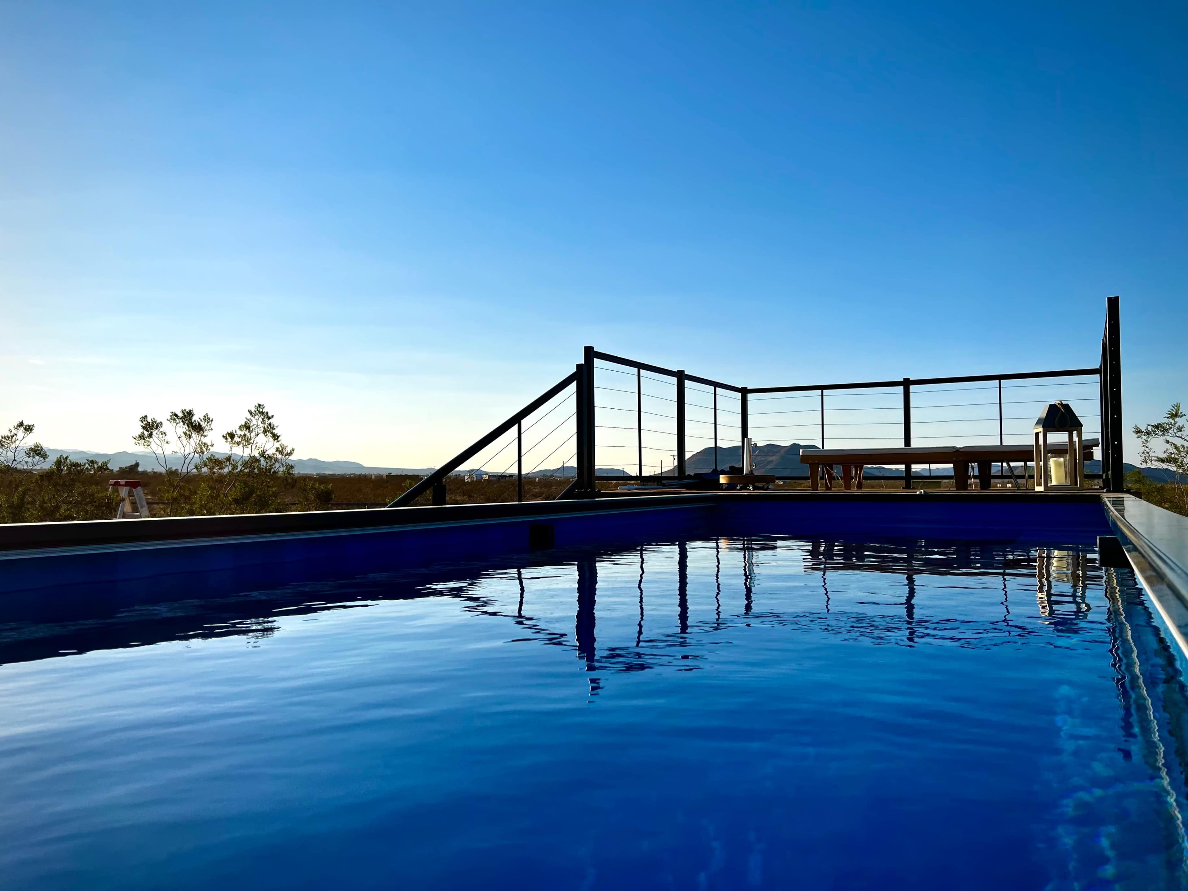 A clear blue pool reflects the sky, with a wooden seating area and mountains visible in the background.