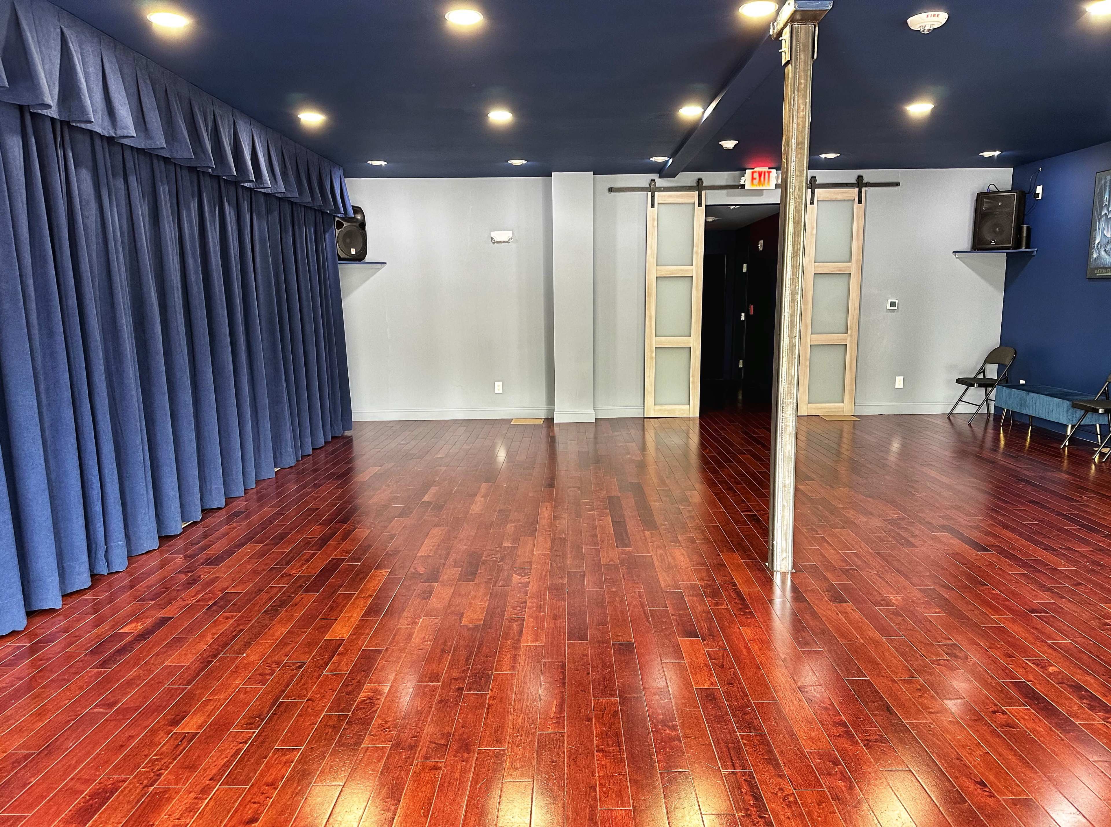 The image depicts an empty dance studio with polished wooden flooring, blue curtains on one side, and two wall-mounted speakers.