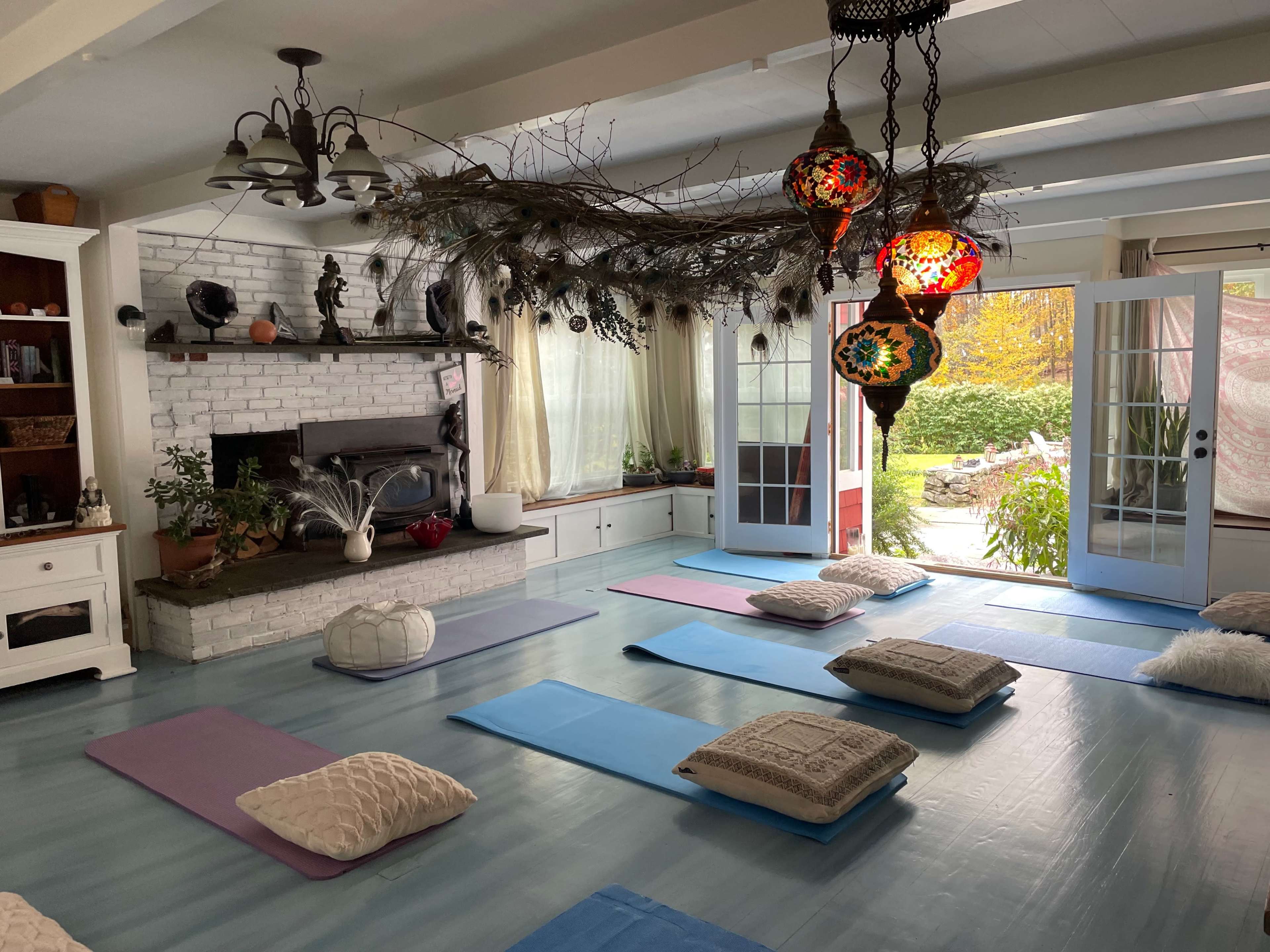 A tranquil yoga studio features colorful mats and cushions arranged neatly beneath a chandelier and alongside a fireplace, with sunlight streaming through large windows.
