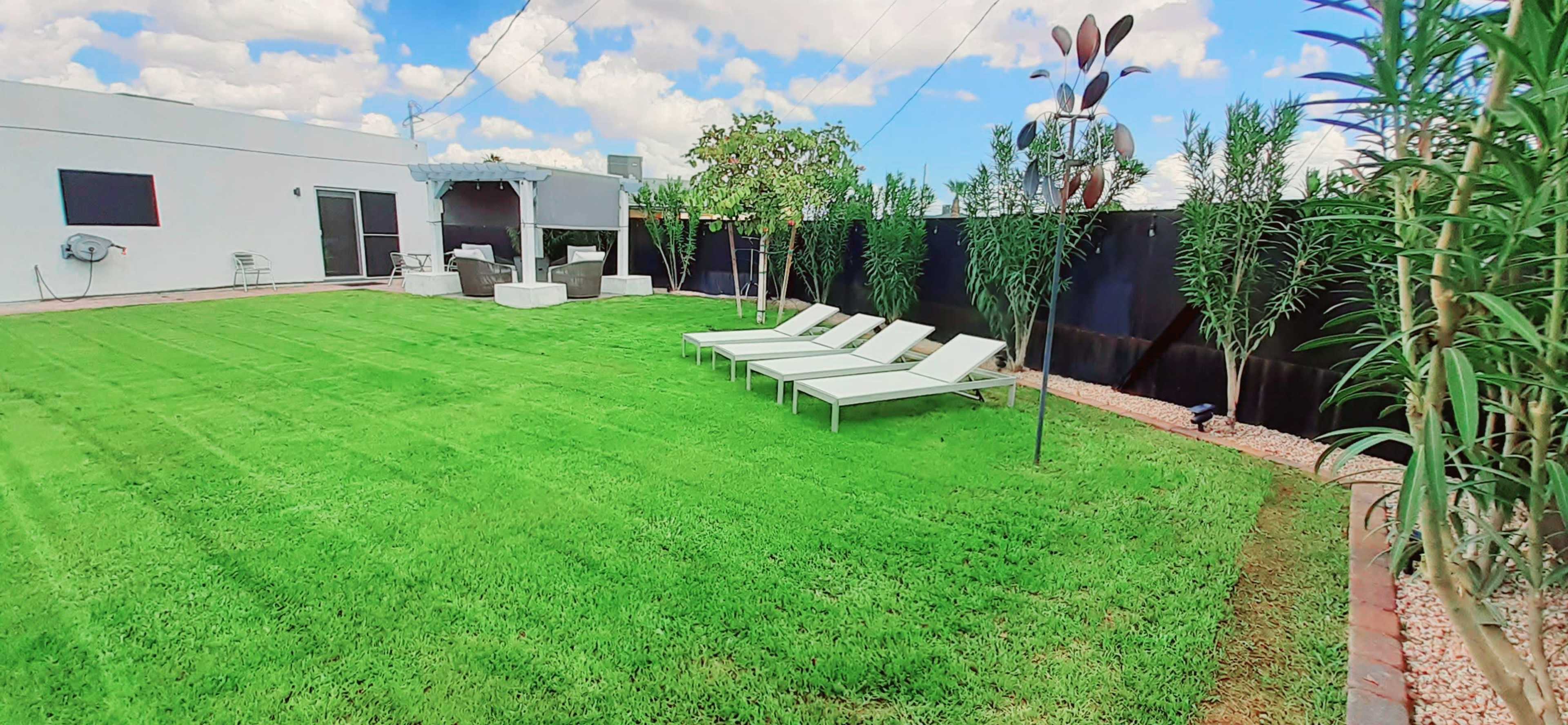 The image shows a well-maintained lawn with three white lounge chairs, a covered patio area, and decorative plants along the perimeter.