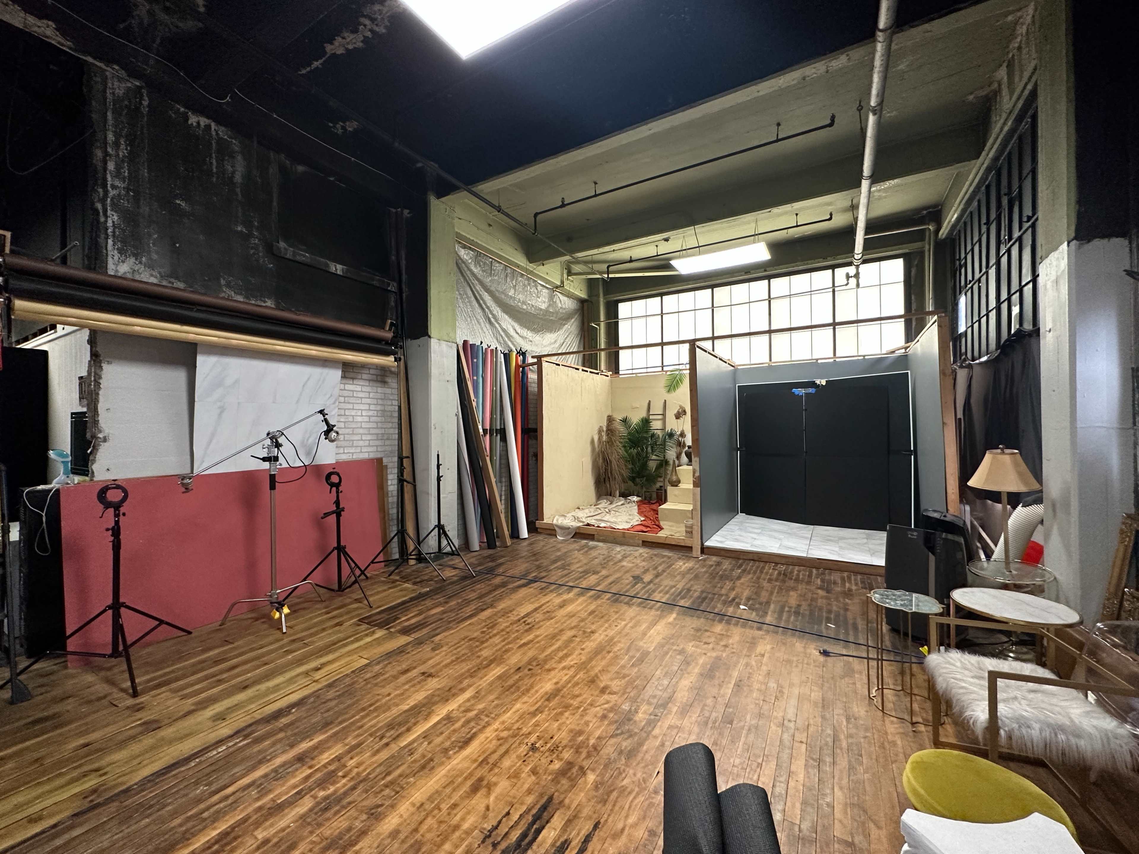 A spacious photography studio with wooden flooring, equipped with lights, a backdrop, and various props including plants and furniture.