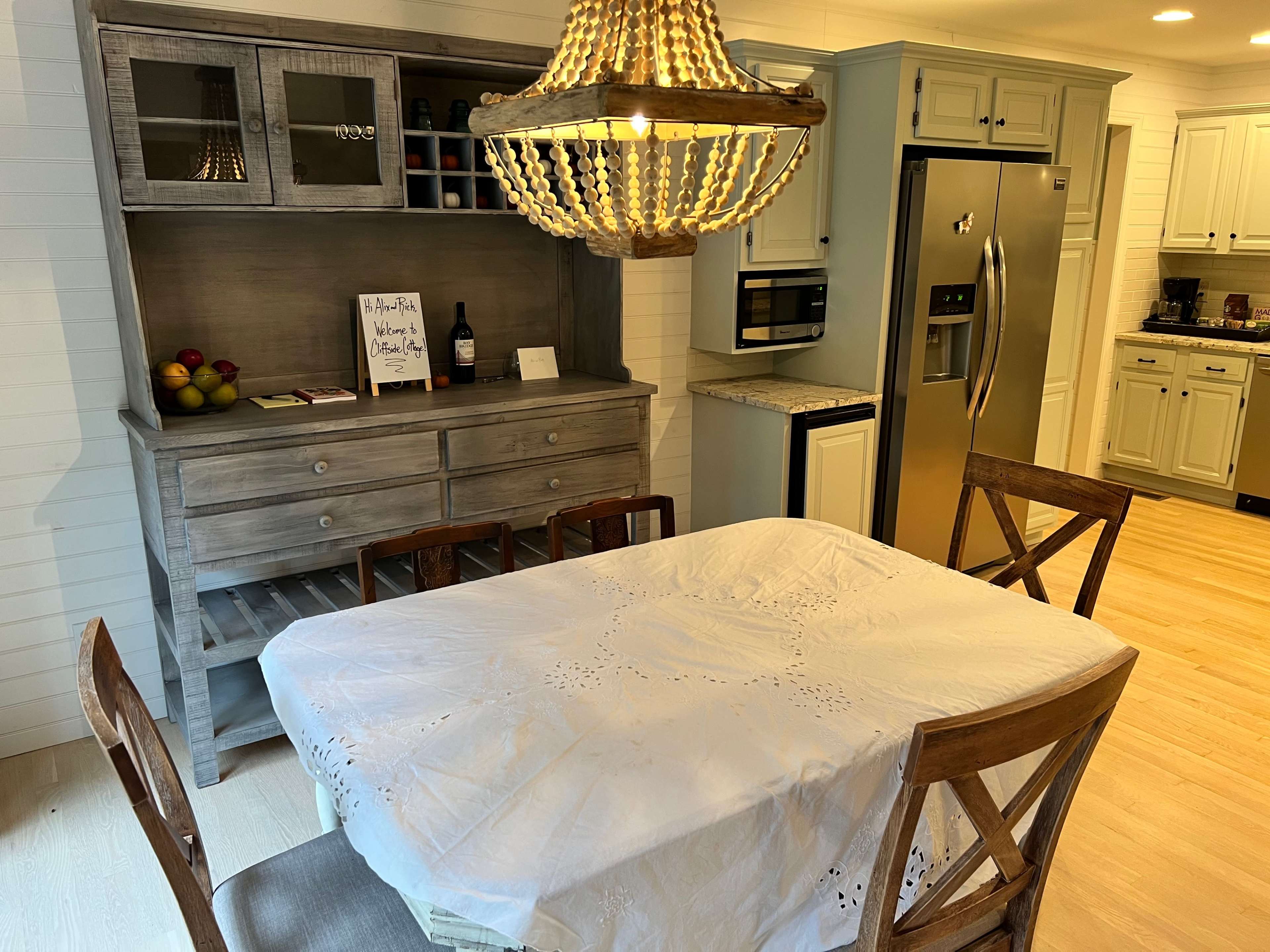 A dining area features a table with a white tablecloth, surrounded by wooden chairs, next to a gray sideboard and a well-equipped kitchen.