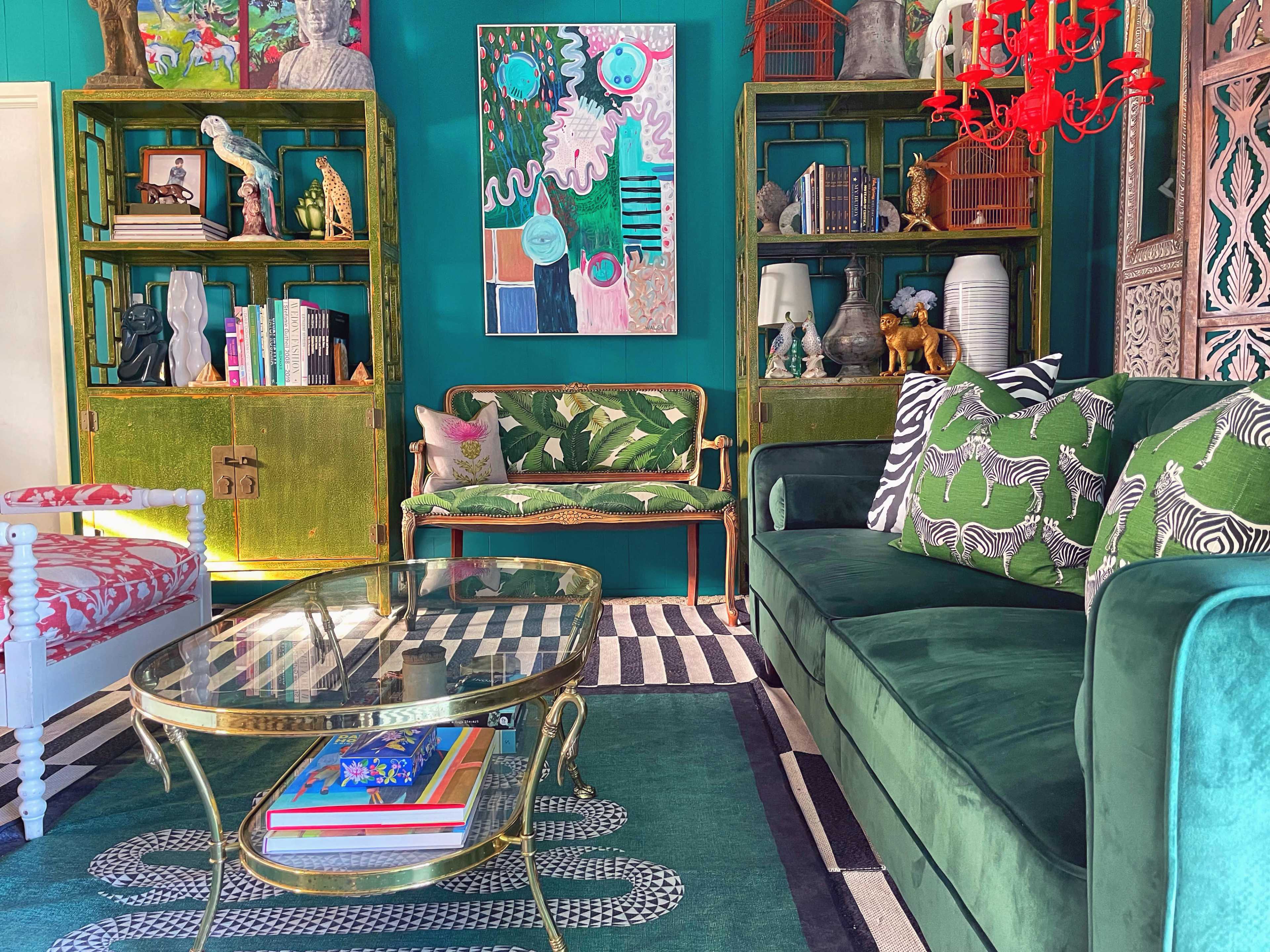 A vibrant living room featuring green walls, a green sofa with patterned pillows, a glass coffee table, and decorated shelves filled with colorful art and objects.
