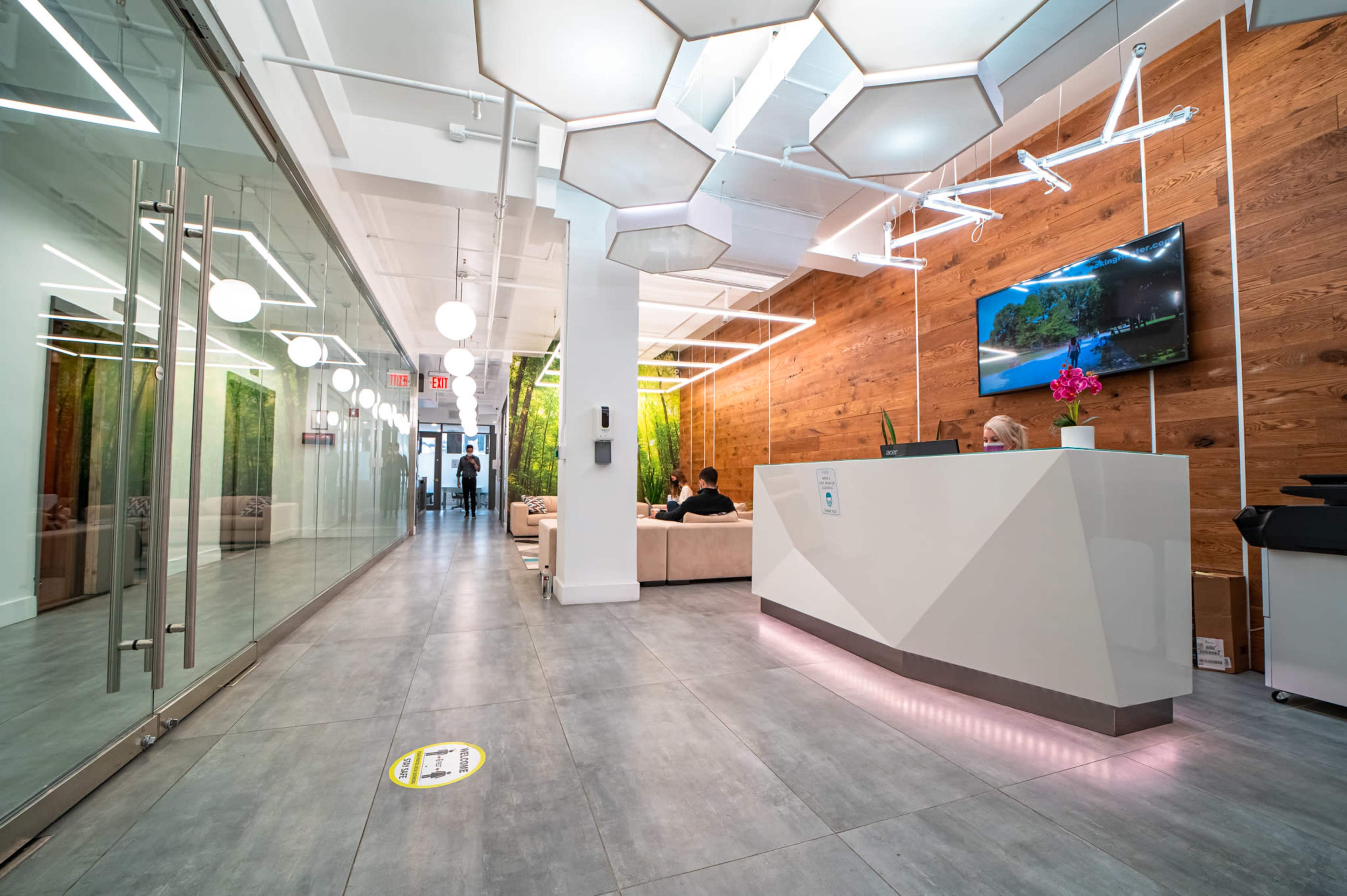 A modern office reception area features a sleek front desk, large glass doors, and a wall adorned with natural wood accents and greenery.