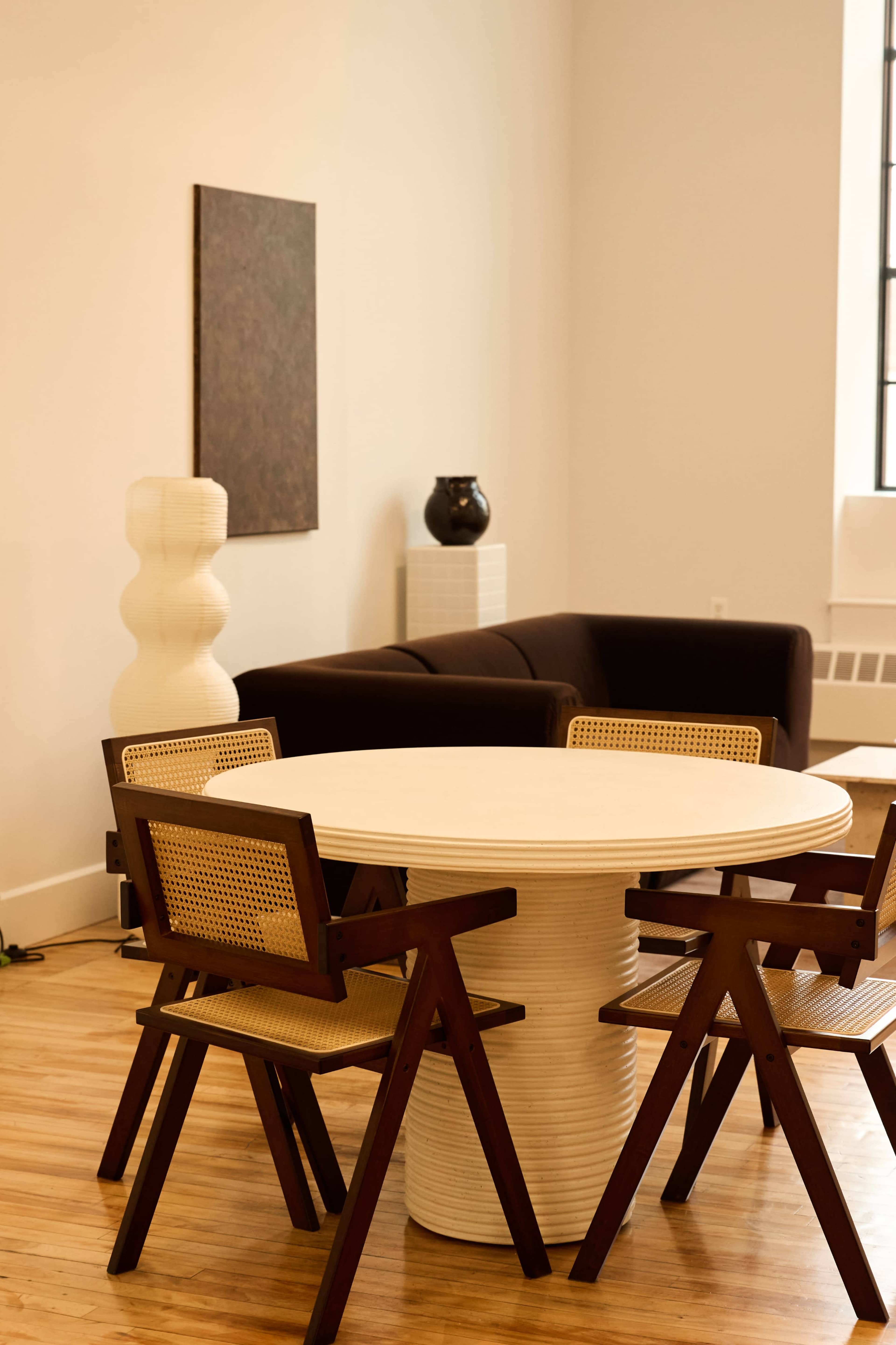 The image features a modern dining area with a round table surrounded by four wooden chairs, complemented by a brown couch and decorative elements in a minimalist space.