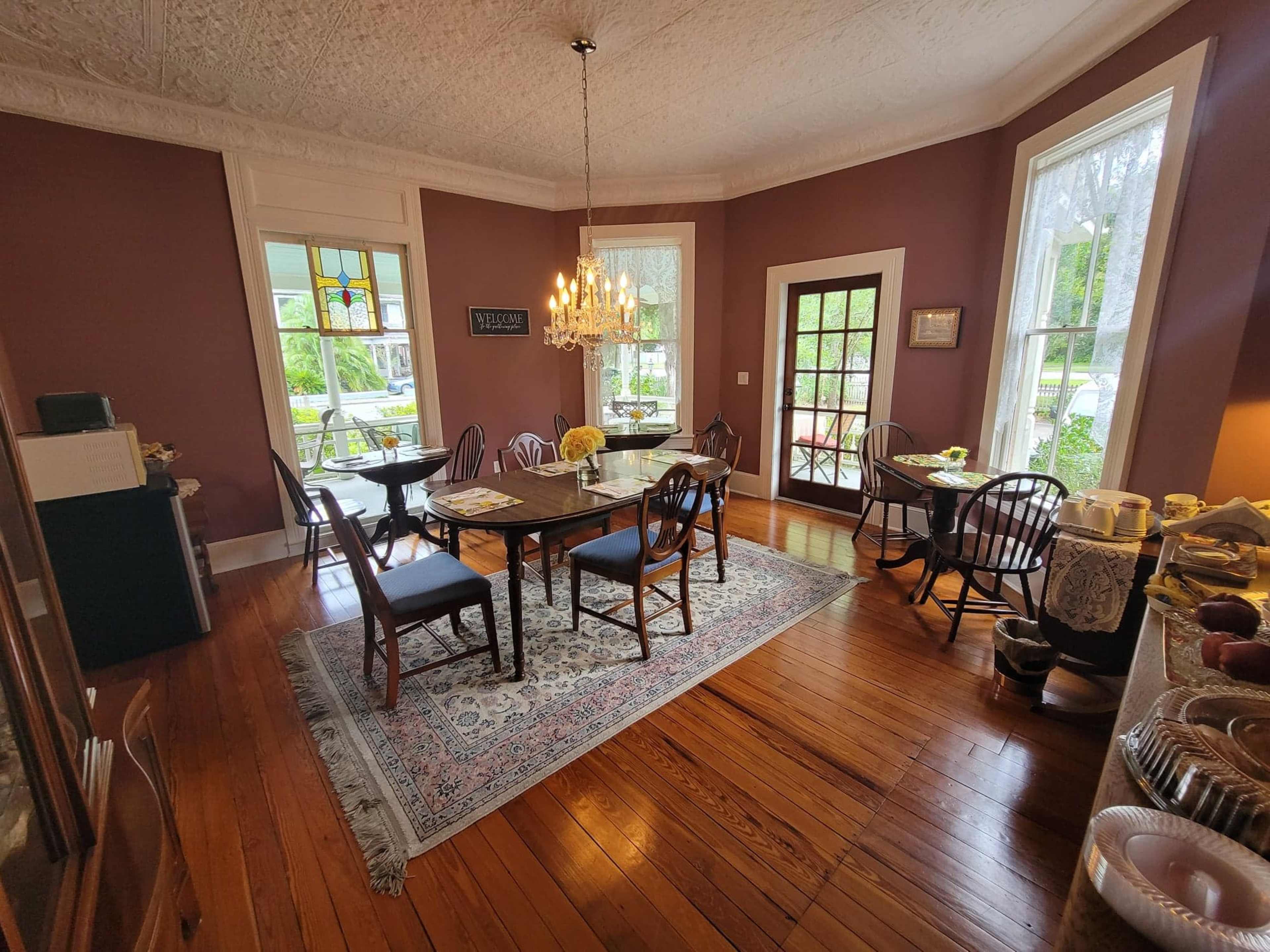 A cozy dining room features a large table surrounded by wooden chairs, with a chandelier hanging above and sunlight streaming through large windows.