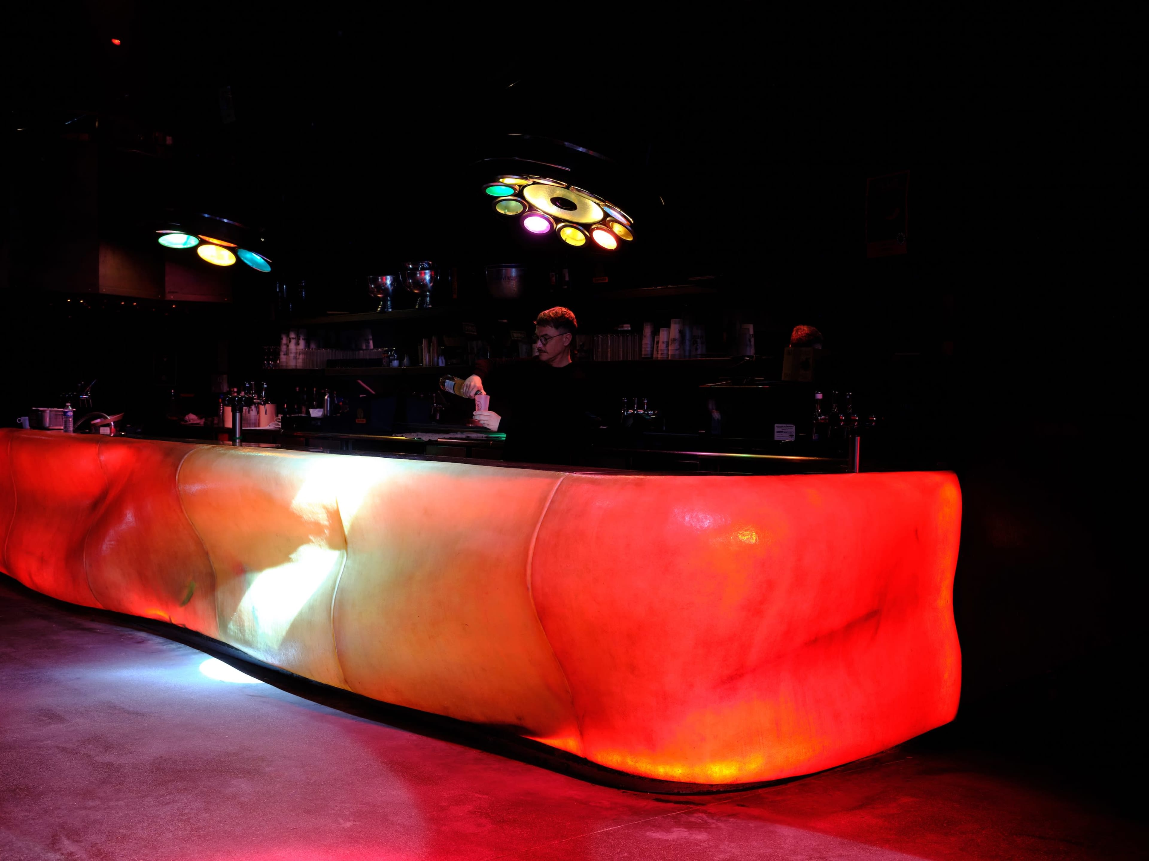 A brightly illuminated bar featuring a sculptural design with vibrant red and orange hues is staffed by a bartender.