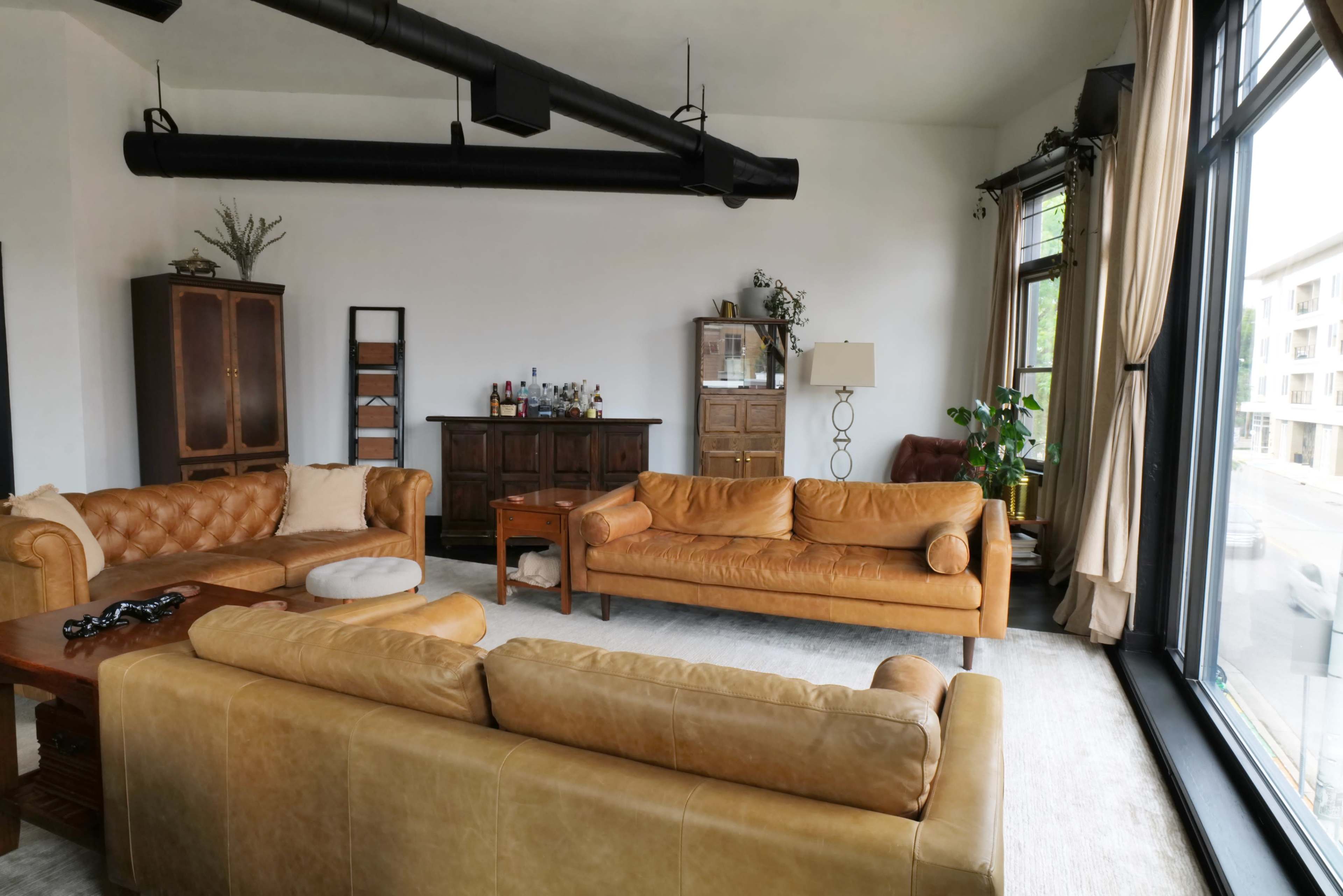 The image shows a spacious living room with leather sofas, a wooden cabinet, and large windows allowing natural light to enter.