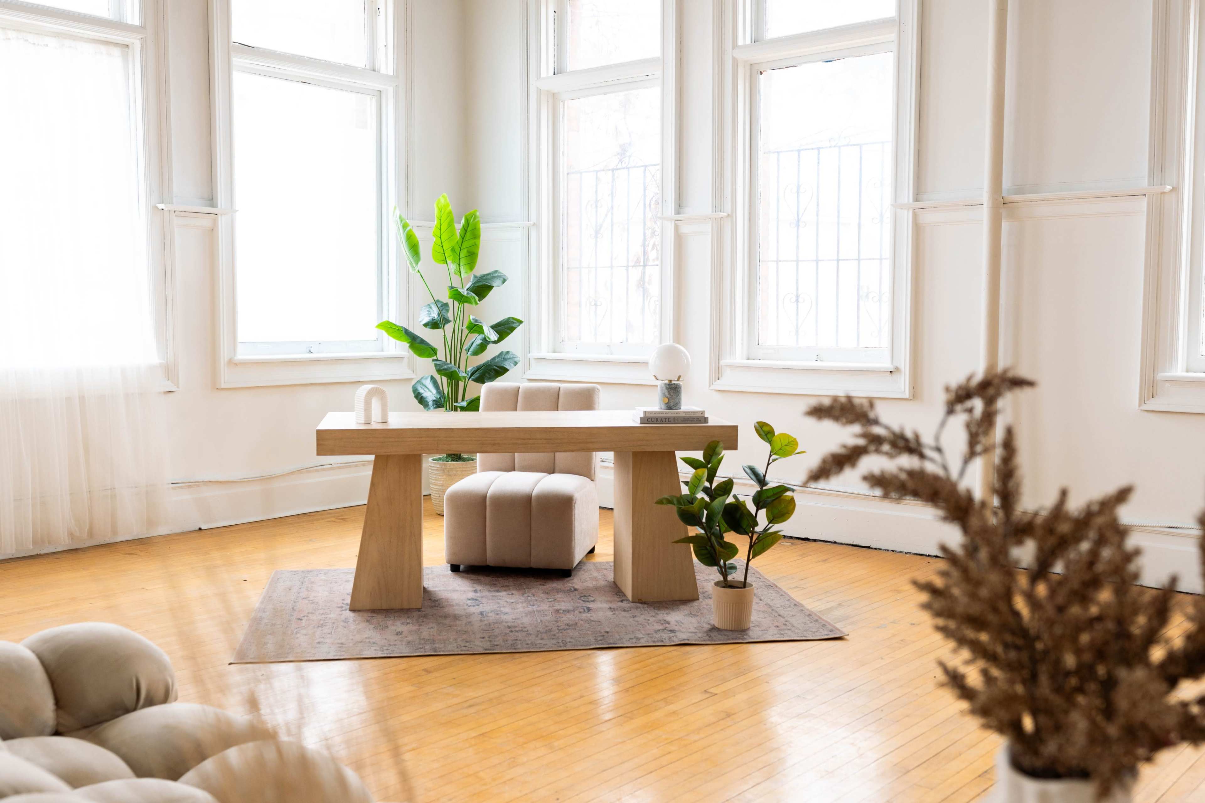 The image depicts a bright and airy room featuring a wooden table, a chair, several potted plants, and large windows allowing natural light to flood the space.