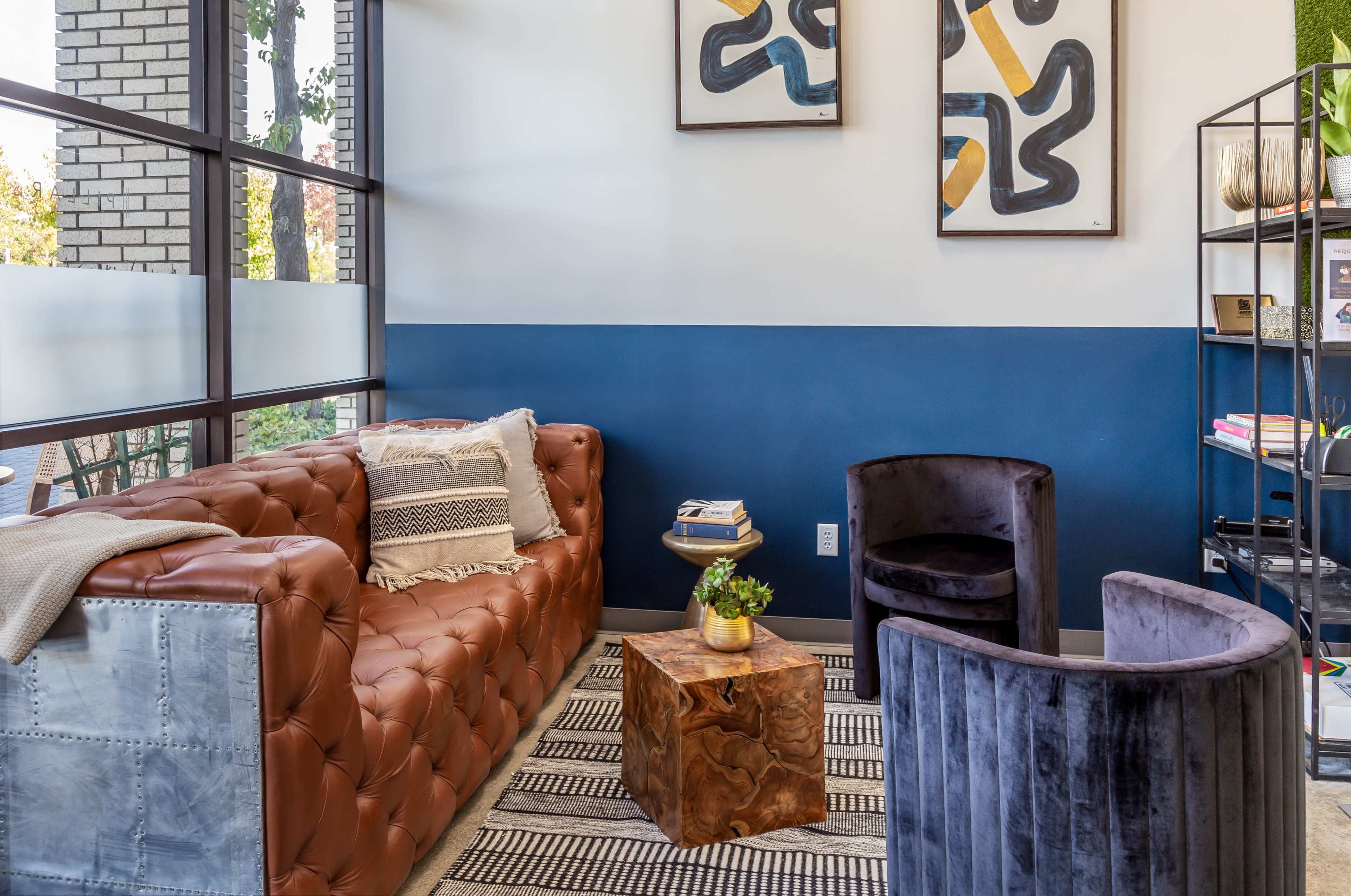 A modern seating area featuring a brown leather sofa, two black velvet chairs, a wooden side table, and abstract artwork on the walls.