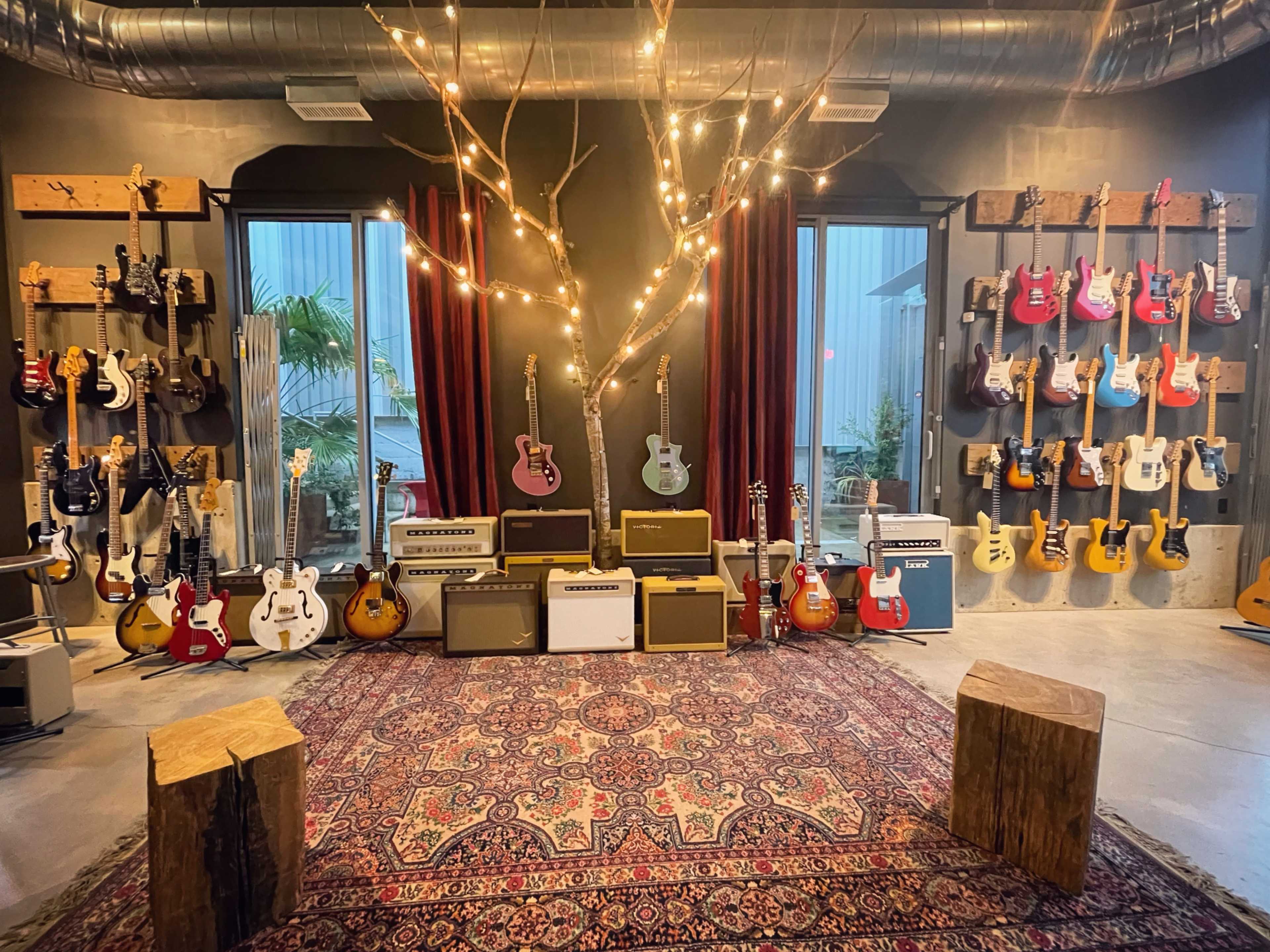 A music studio with various guitars displayed on the walls, amplifier stacks in the center, and wooden stools on a patterned rug.