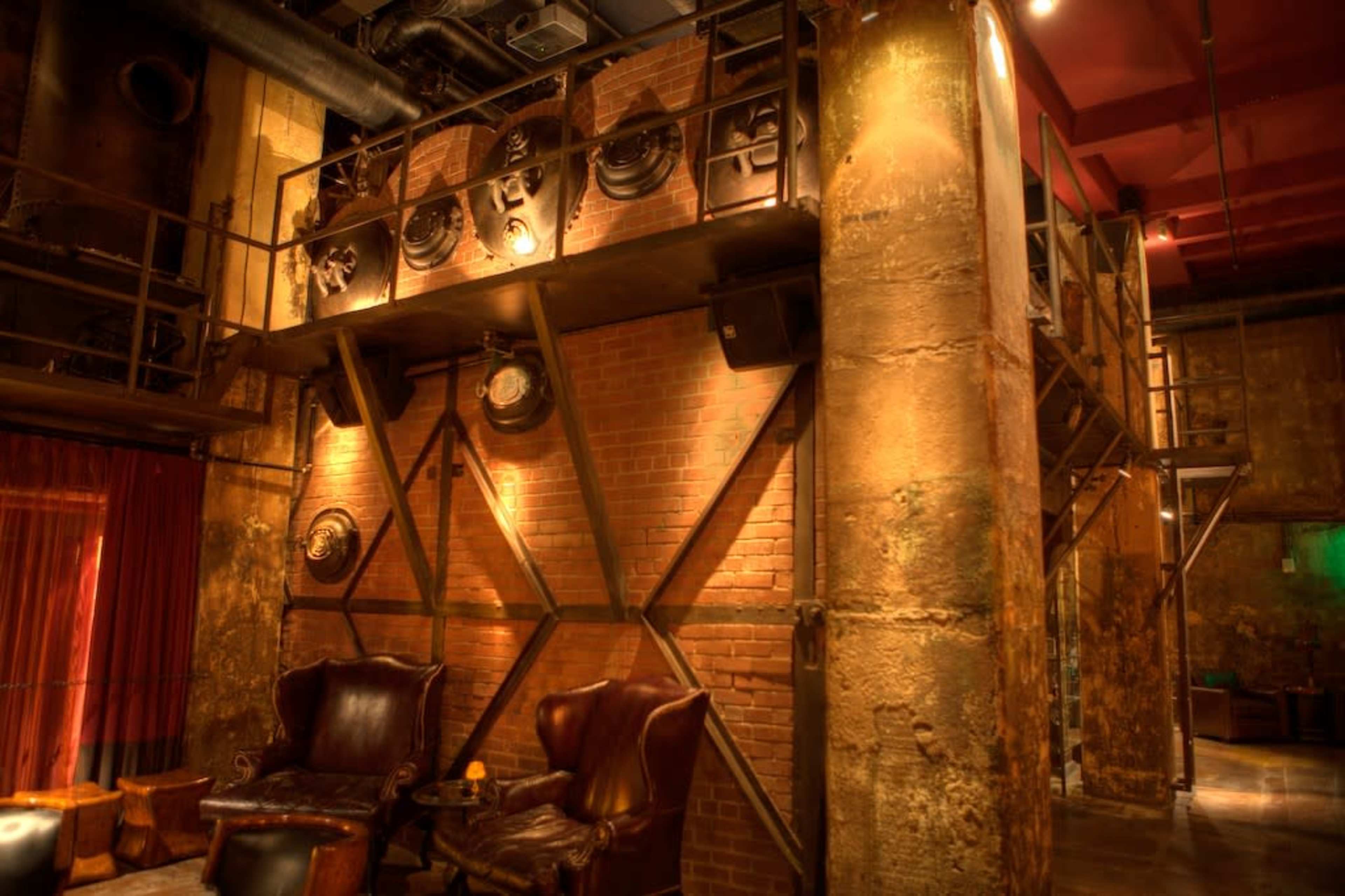 The image shows a dimly lit industrial-style bar interior with exposed brick walls, metal accents, and leather seating.