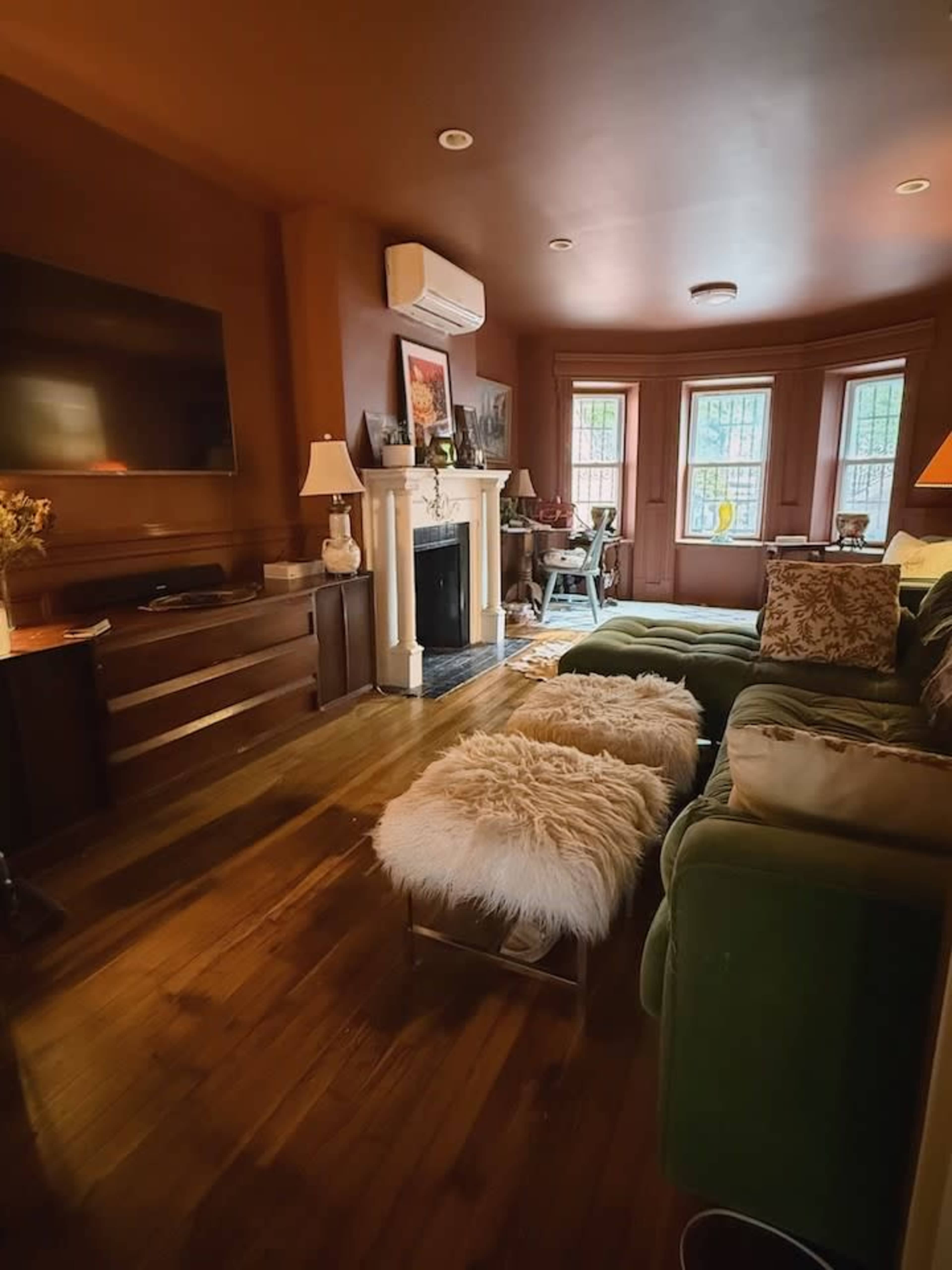The image shows a cozy living room with a fireplace, a large television, and a green couch facing a set of windows.