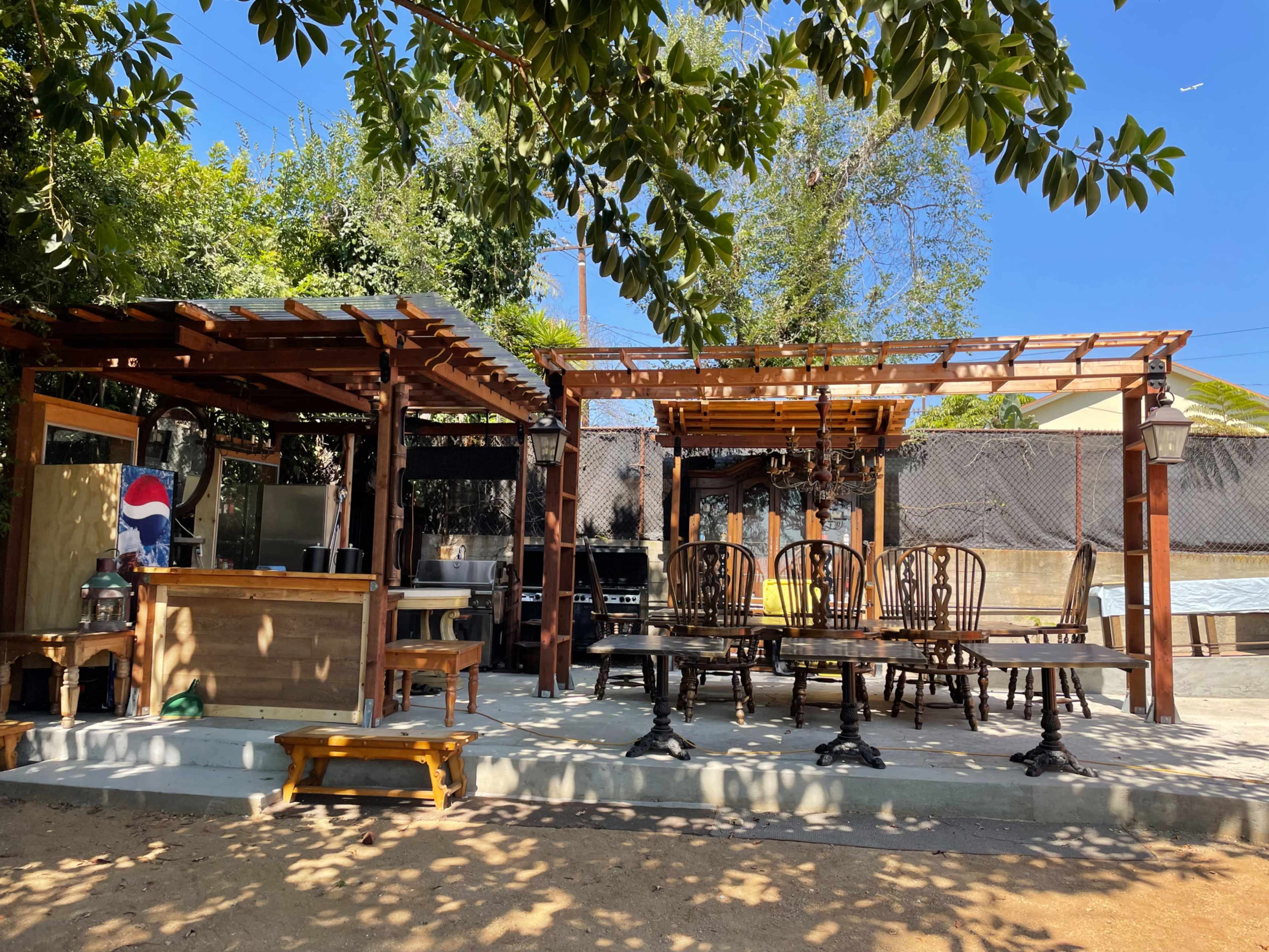 The image shows an outdoor seating area with wooden tables and chairs, a bar structure, and various trees and plants in the background.
