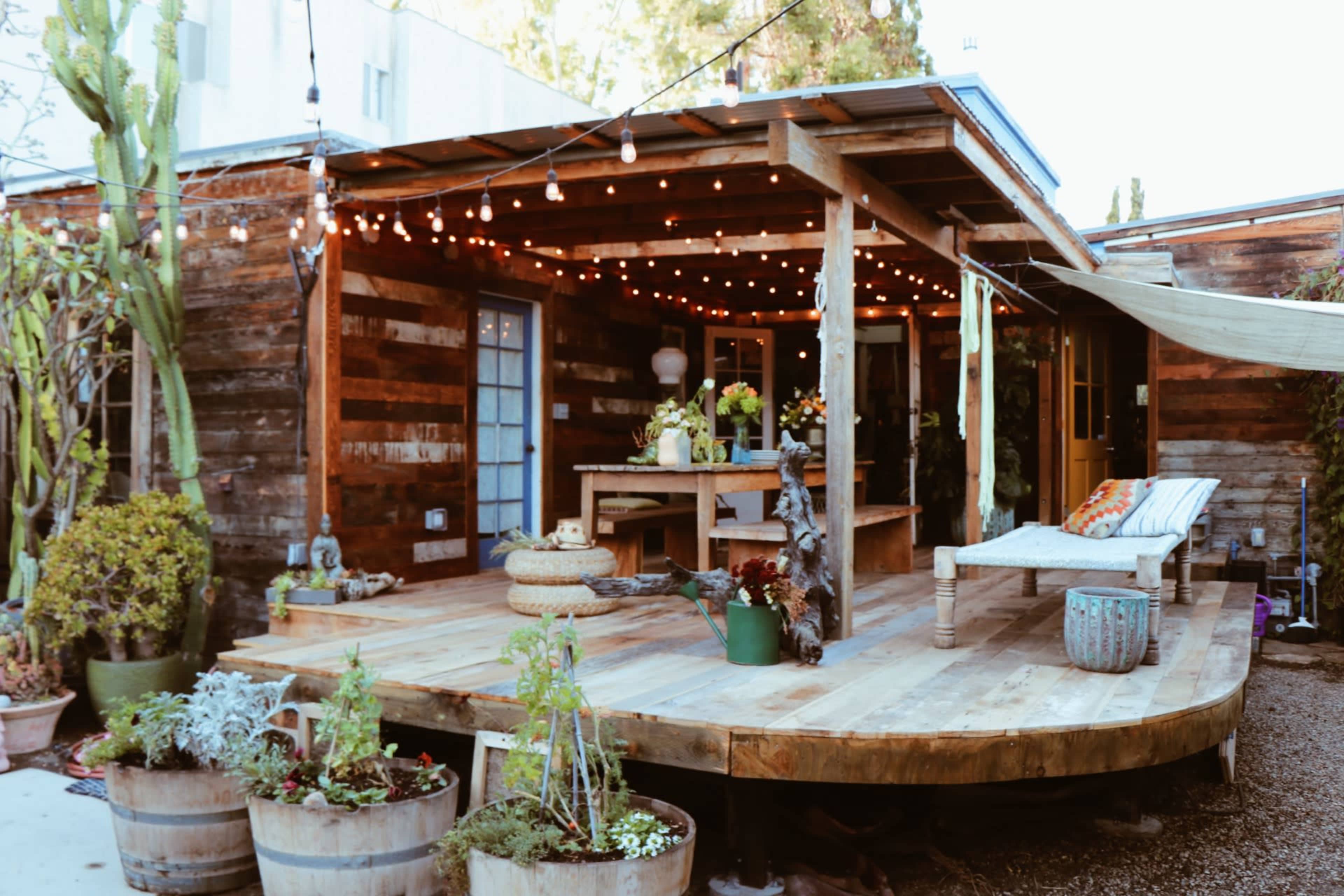 The image shows a rustic outdoor patio area with wooden decking, potted plants, and string lights illuminating the space.