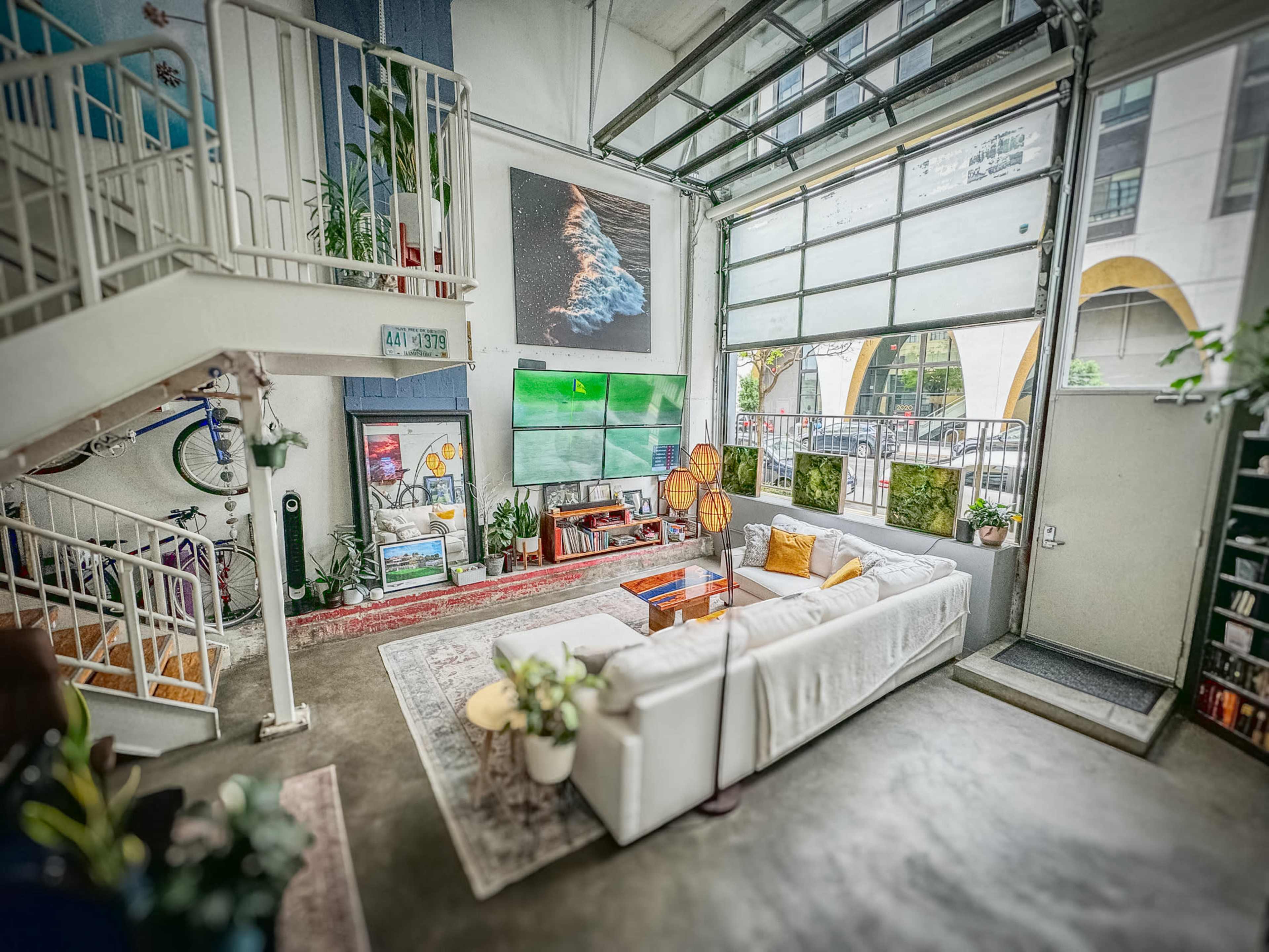 The image shows a spacious, modern living room with large windows, a white sectional sofa, a coffee table, and various artworks on the walls, alongside a staircase and bike in the corner.