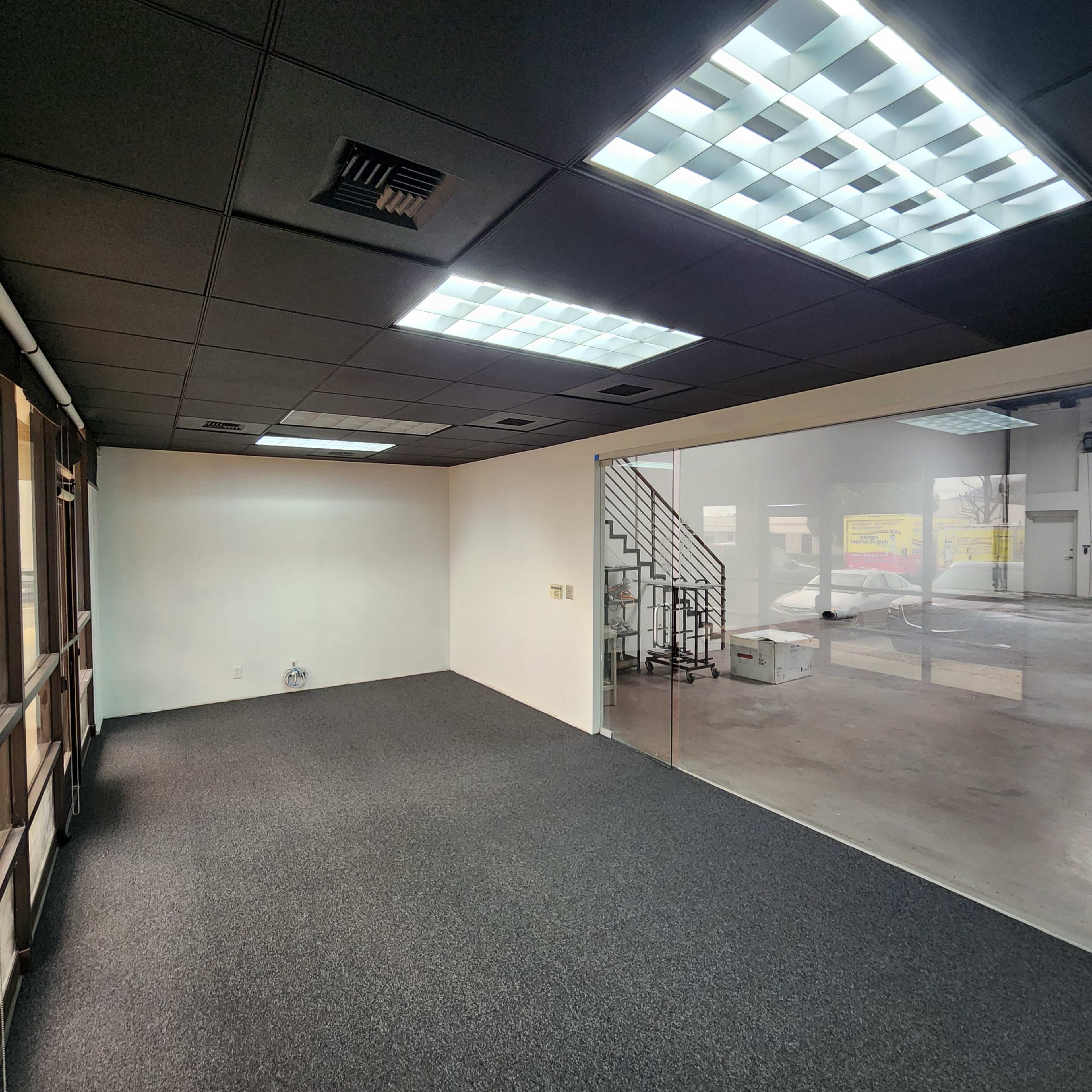 An empty office space with black carpeting, white walls, and fluorescent ceiling lights, featuring a glass partition that provides a view of an adjacent area.