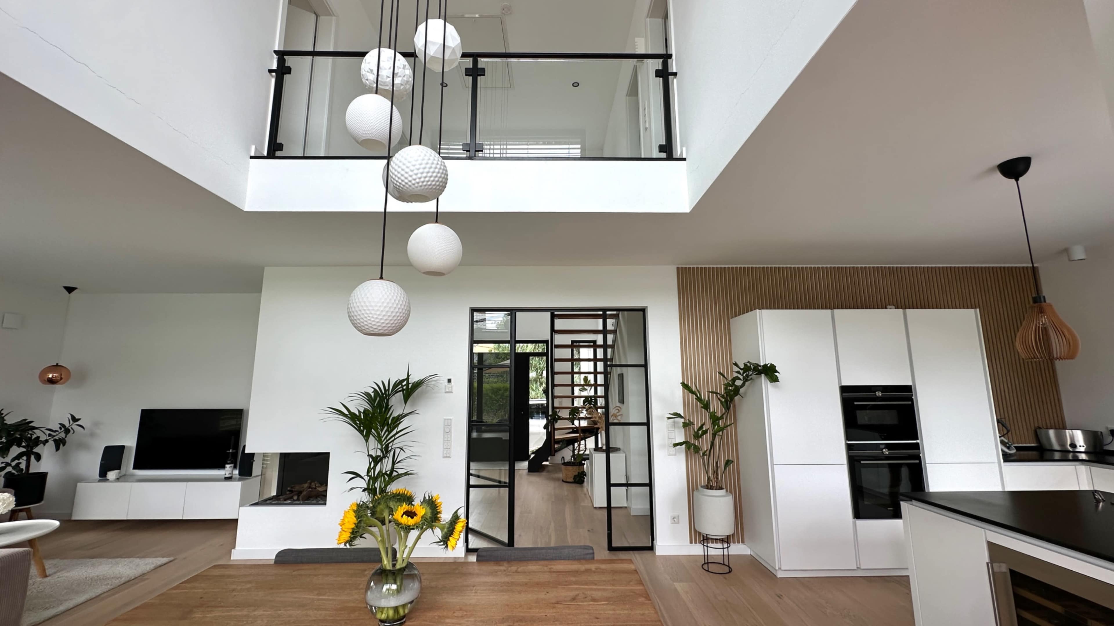 The image shows a modern, open-concept living space featuring a dining table with a vase of sunflowers, a staircase leading to an upper level, and large windows allowing natural light.