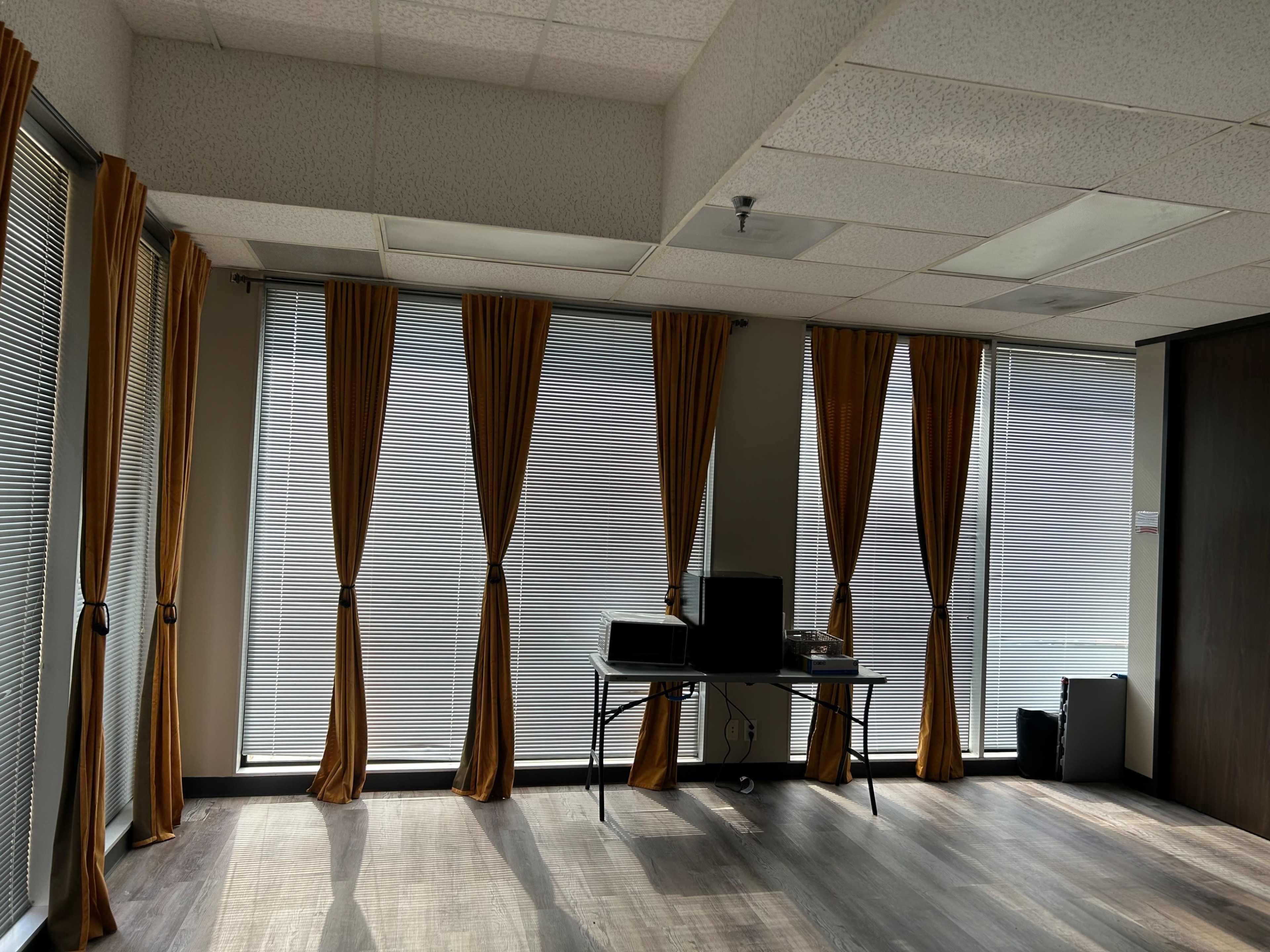 The image shows a sparsely furnished office space with large windows covered by brown curtains and a desk with a computer in front of one window.
