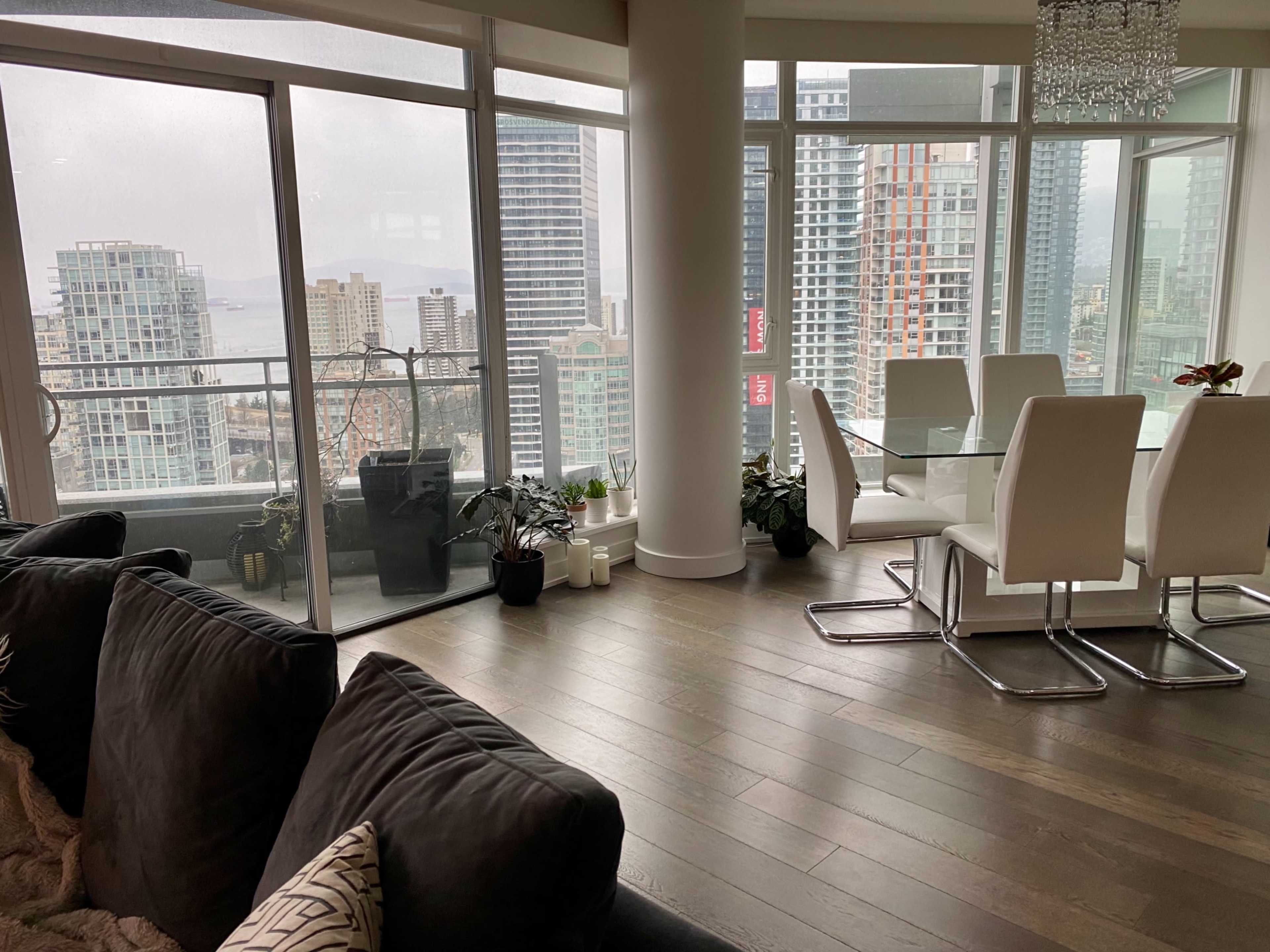 The image shows a modern apartment interior with large windows overlooking a city skyline, featuring a dining area and a cozy seating arrangement.