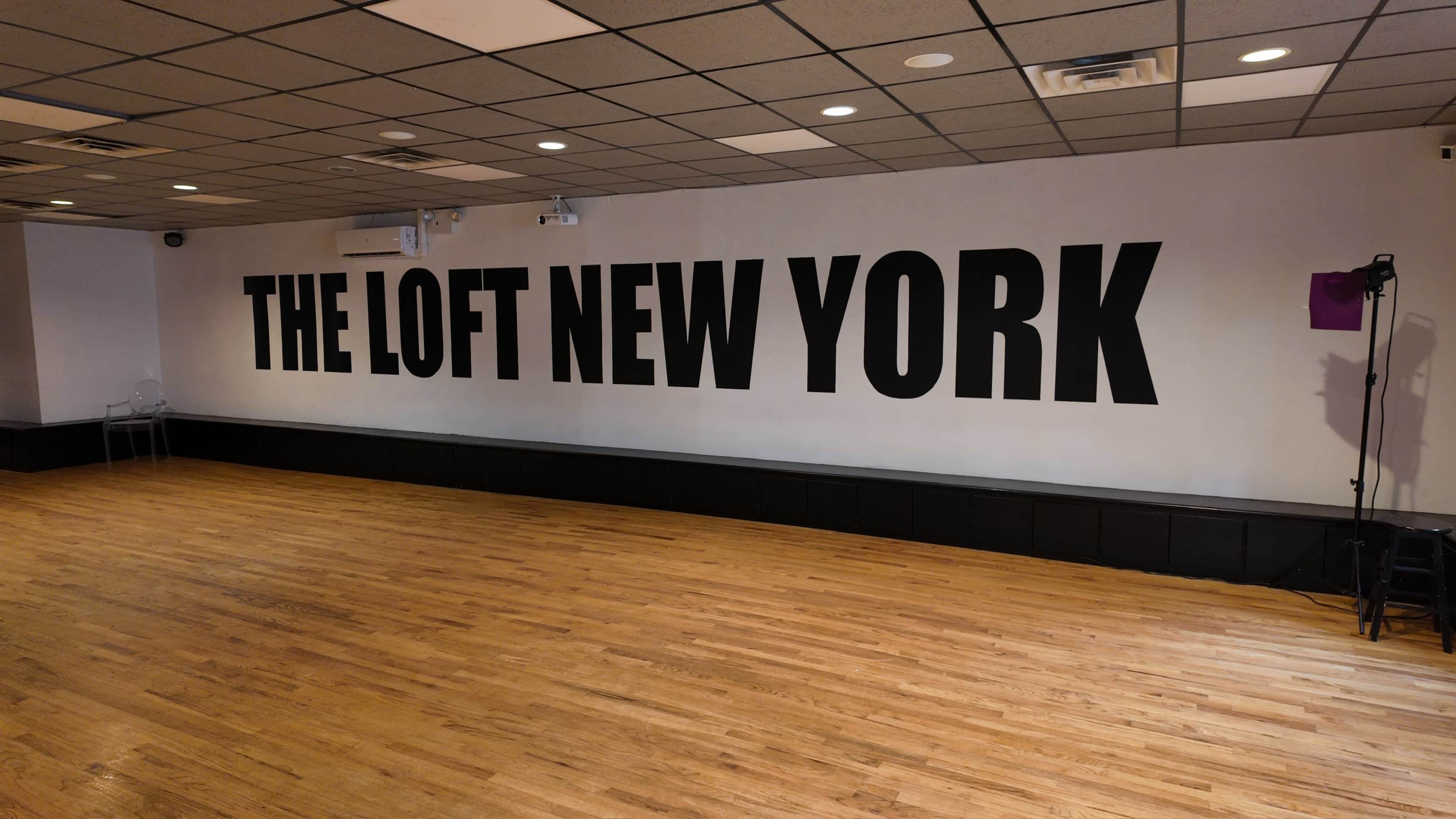 The image shows a spacious room with wooden flooring and a prominent wall featuring the text "THE LOFT NEW YORK" in large, bold letters.