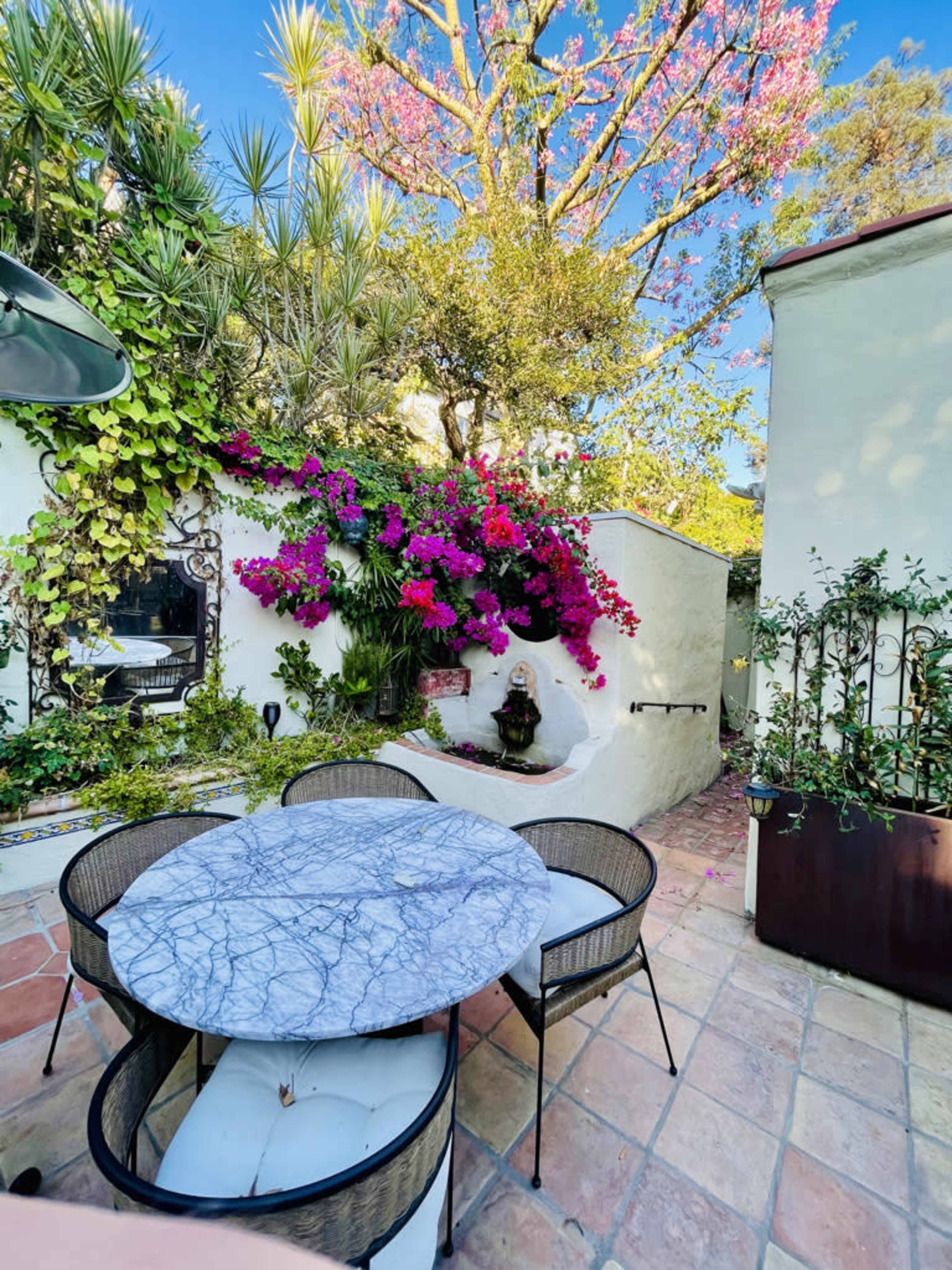 A vibrant garden patio features a round marble table surrounded by black wicker chairs, with lush flowering plants and a water fountain in the background.