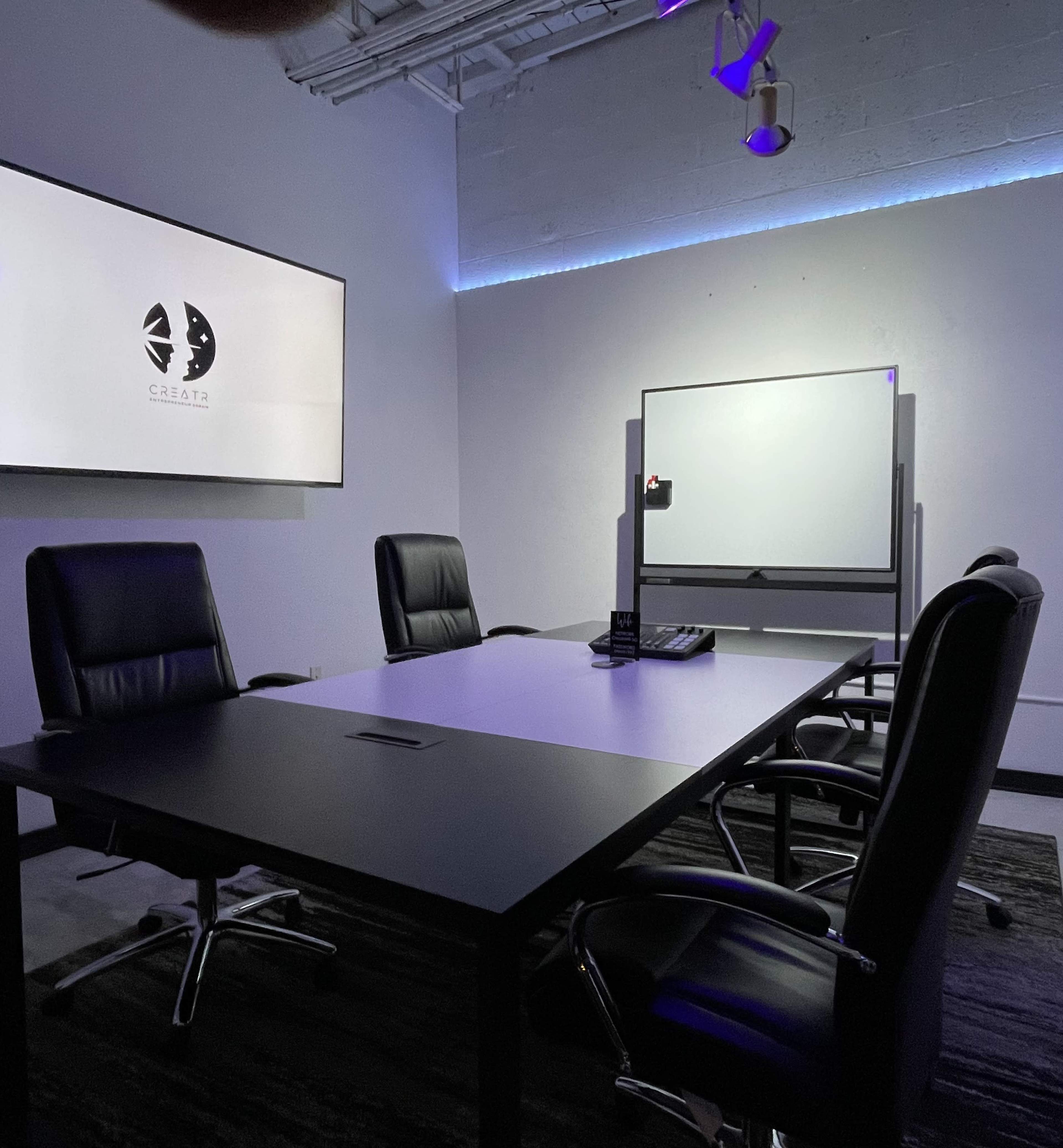 A modern conference room features a large table surrounded by black leather chairs, with a wall-mounted screen and a whiteboard in the background.