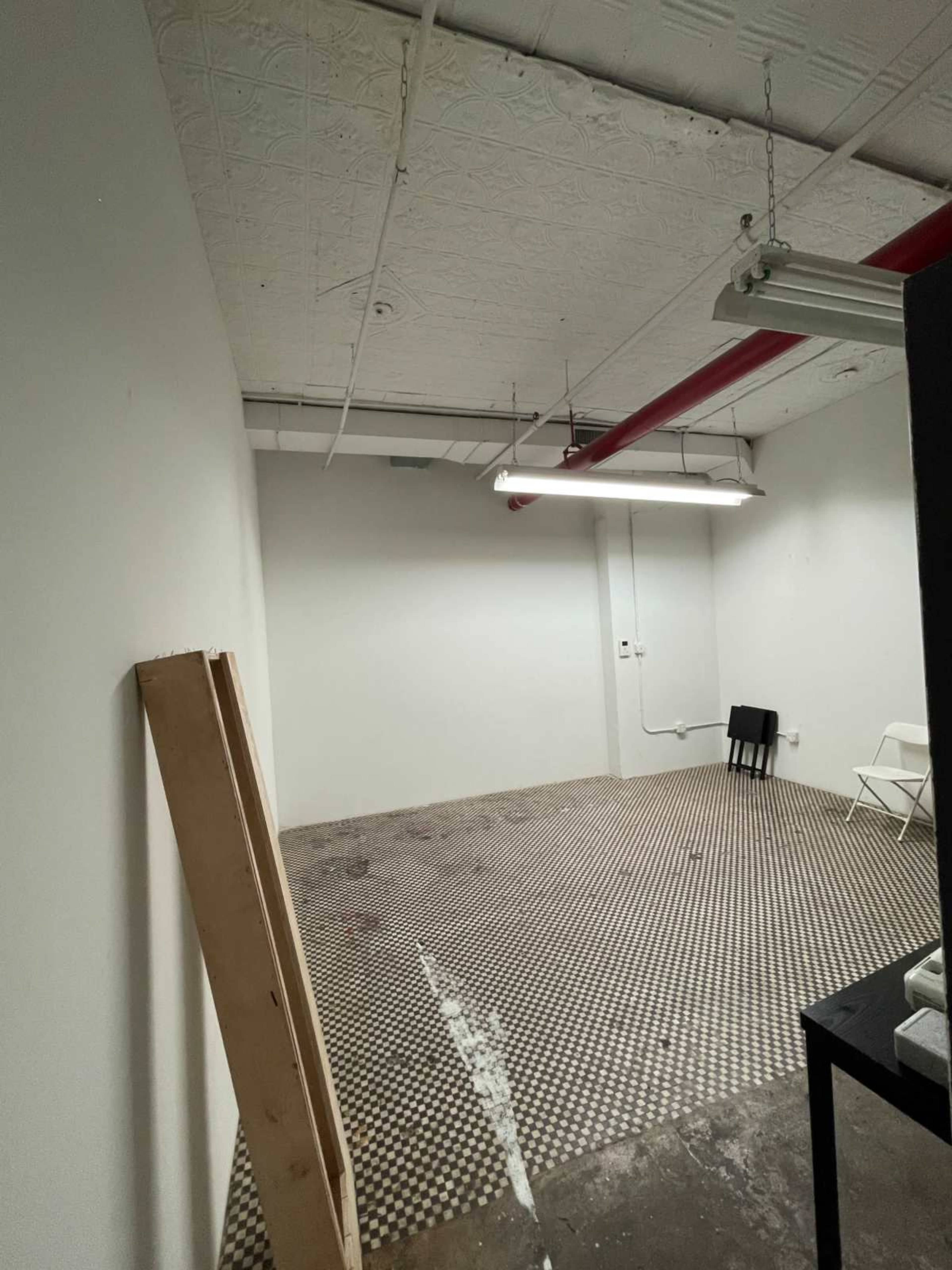 The image shows an empty room with checkerboard flooring, a white ceiling, and minimal furnishings, including a chair and some wooden planks.