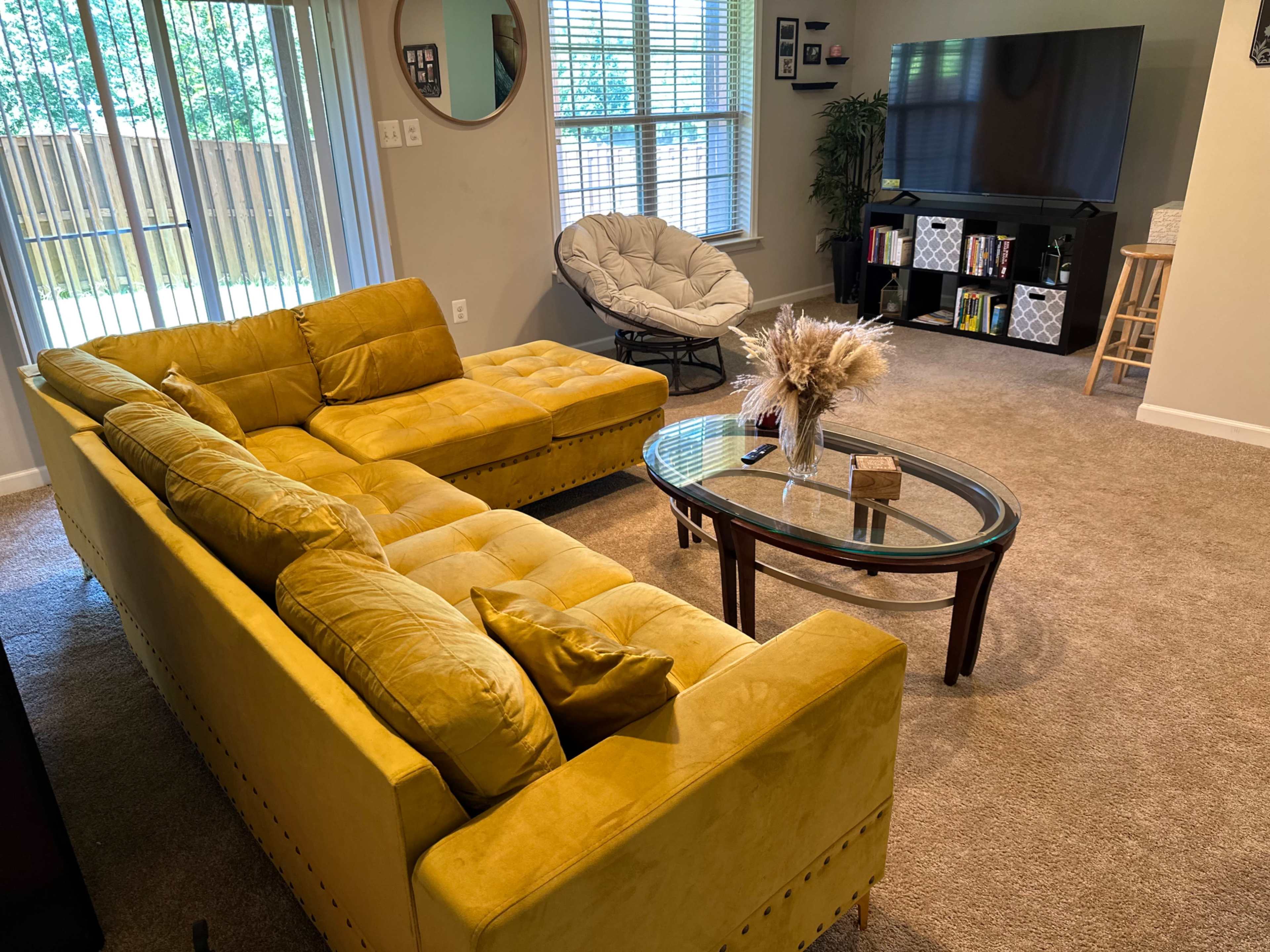 A living room with a yellow sectional sofa, a glass coffee table in the center, and a TV stand with bookshelves against the wall.
