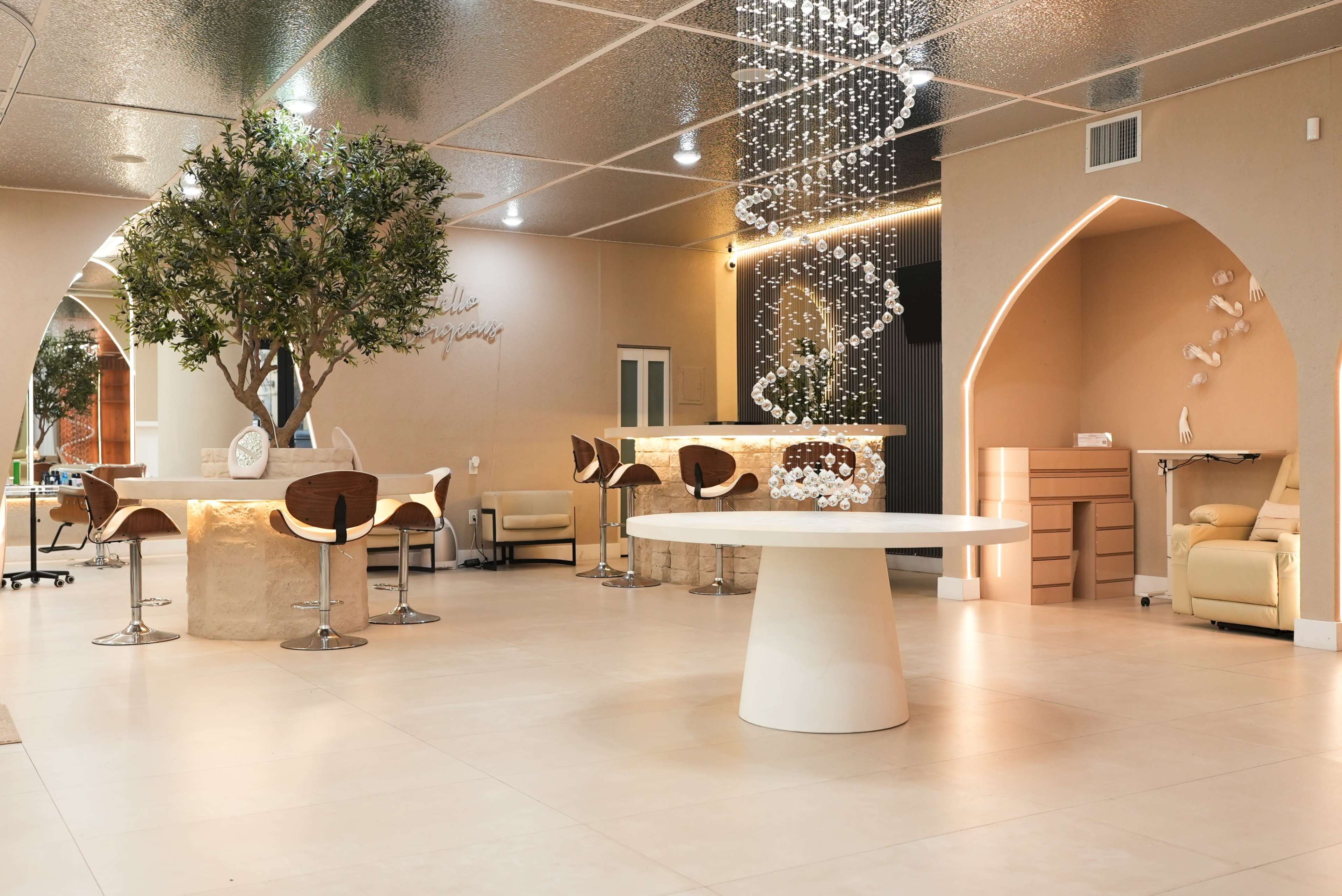 The image shows a modern interior of a spa or wellness center featuring a central round table, bar seating, a decorative tree, and a stylish light fixture.