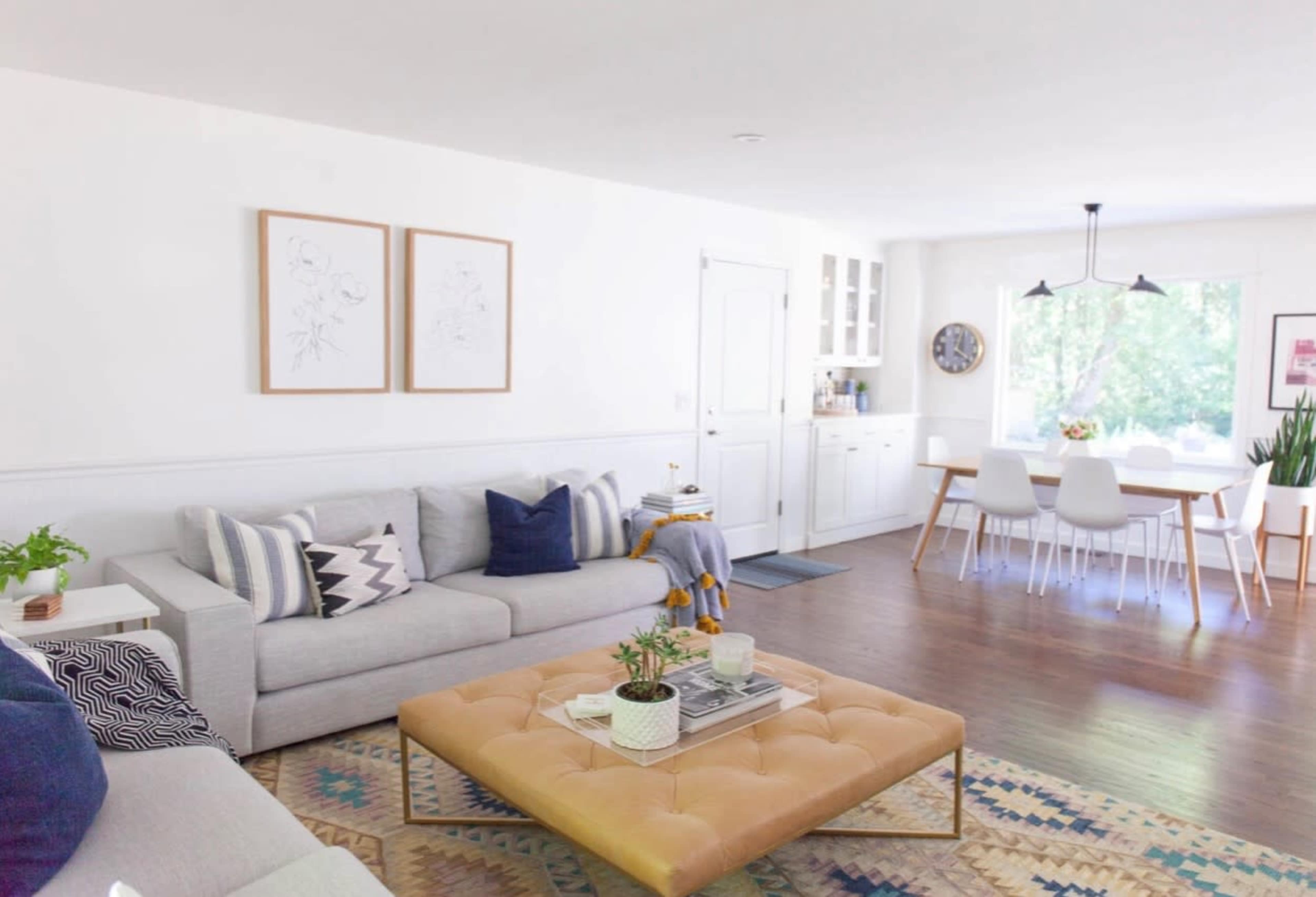 A bright living room with a gray sofa, a large tan ottoman, a patterned rug, and a dining area with white chairs and a large window.