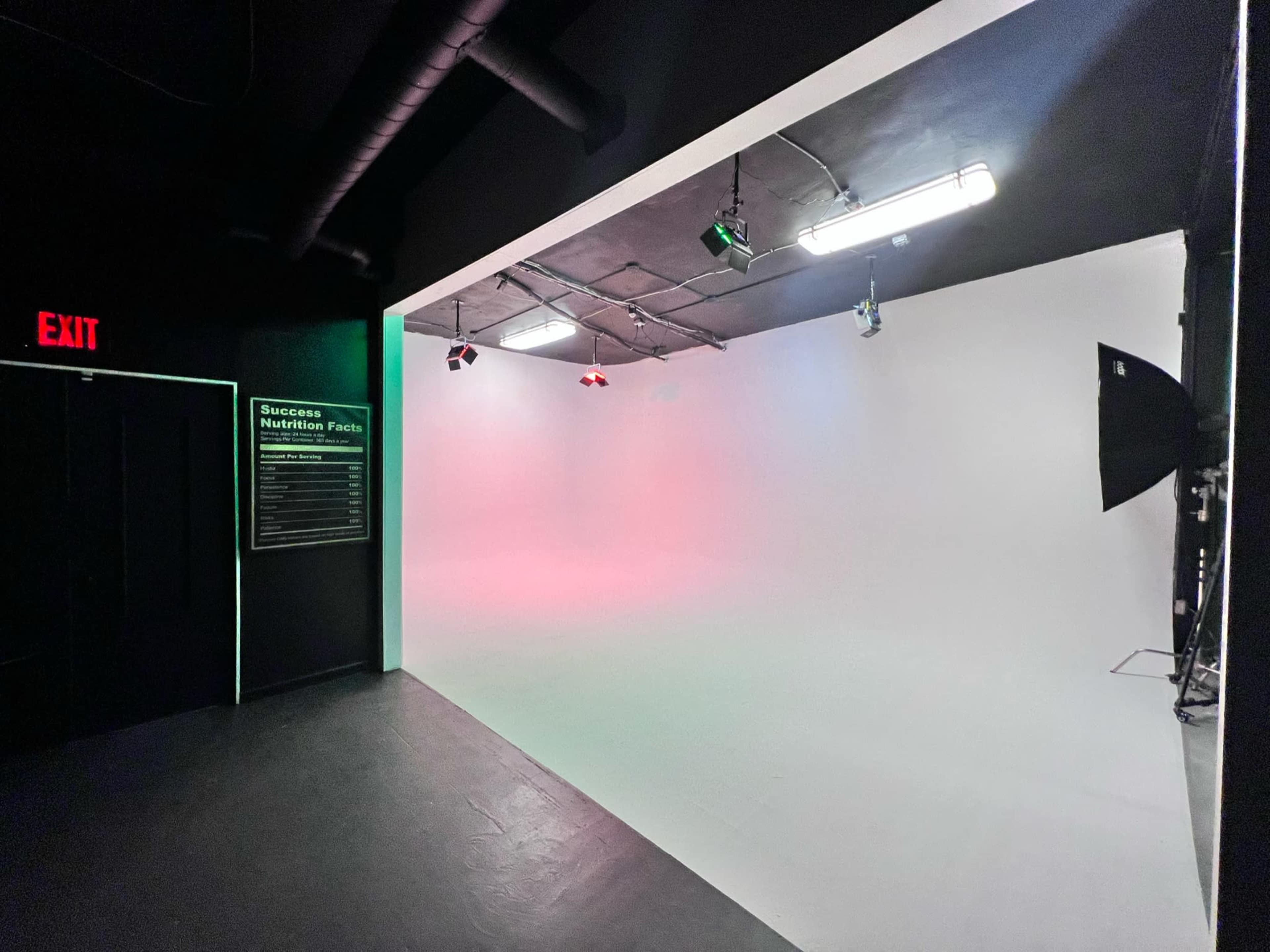 A studio space with a plain white backdrop, lighting fixtures on the ceiling, and an exit sign on the left wall.