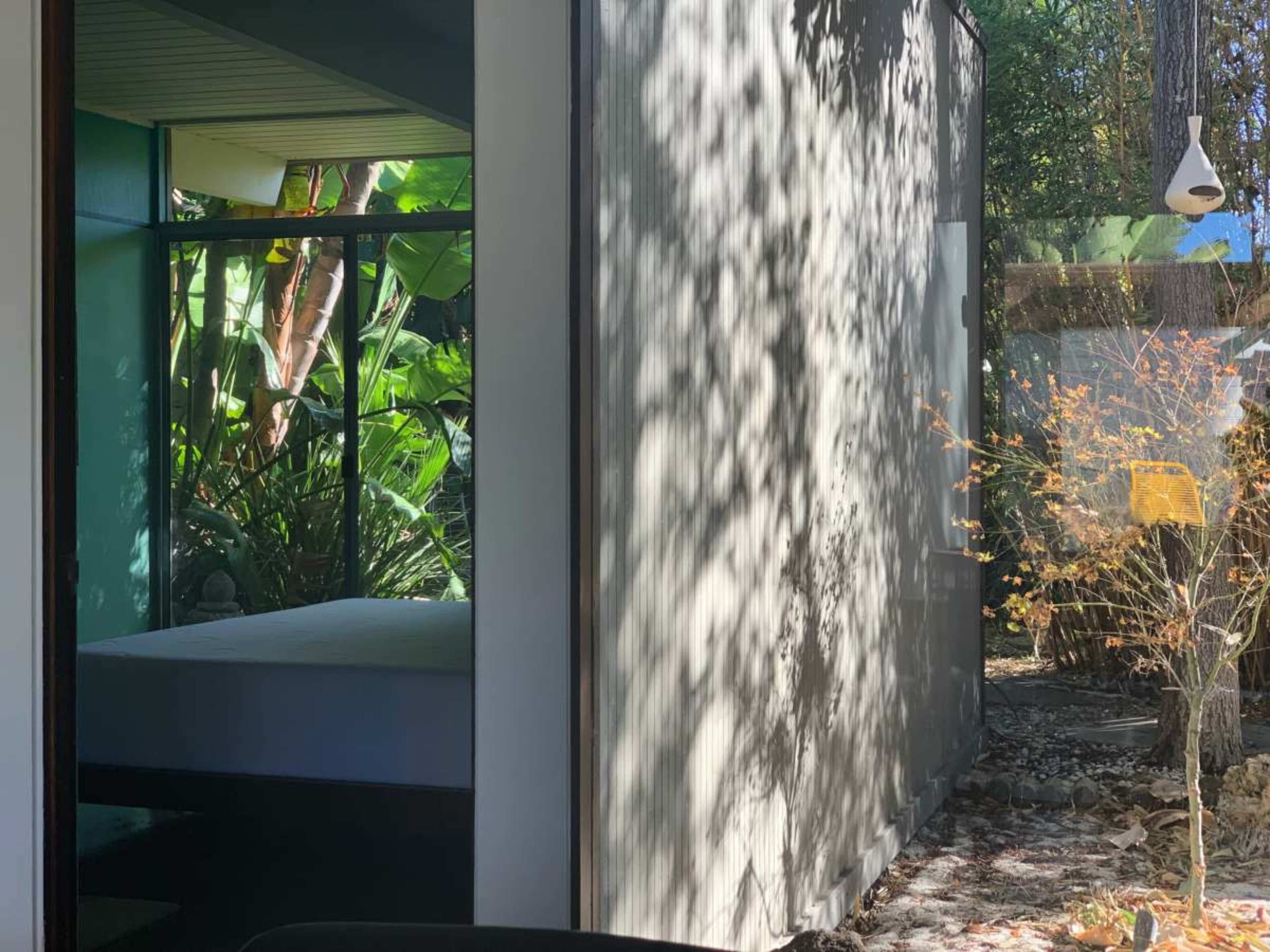 The image shows a view from inside a modern structure, featuring a bed and a bright window that looks out onto a garden with tropical plants.