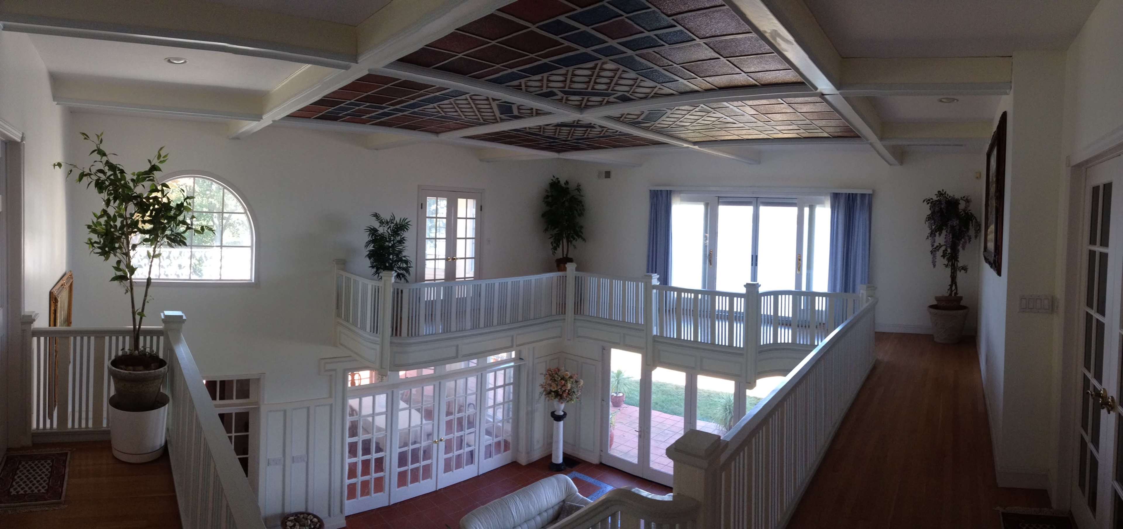 The image shows a spacious interior with a second-floor balcony overlooking a living area, featuring large windows and decorative ceiling tiles.