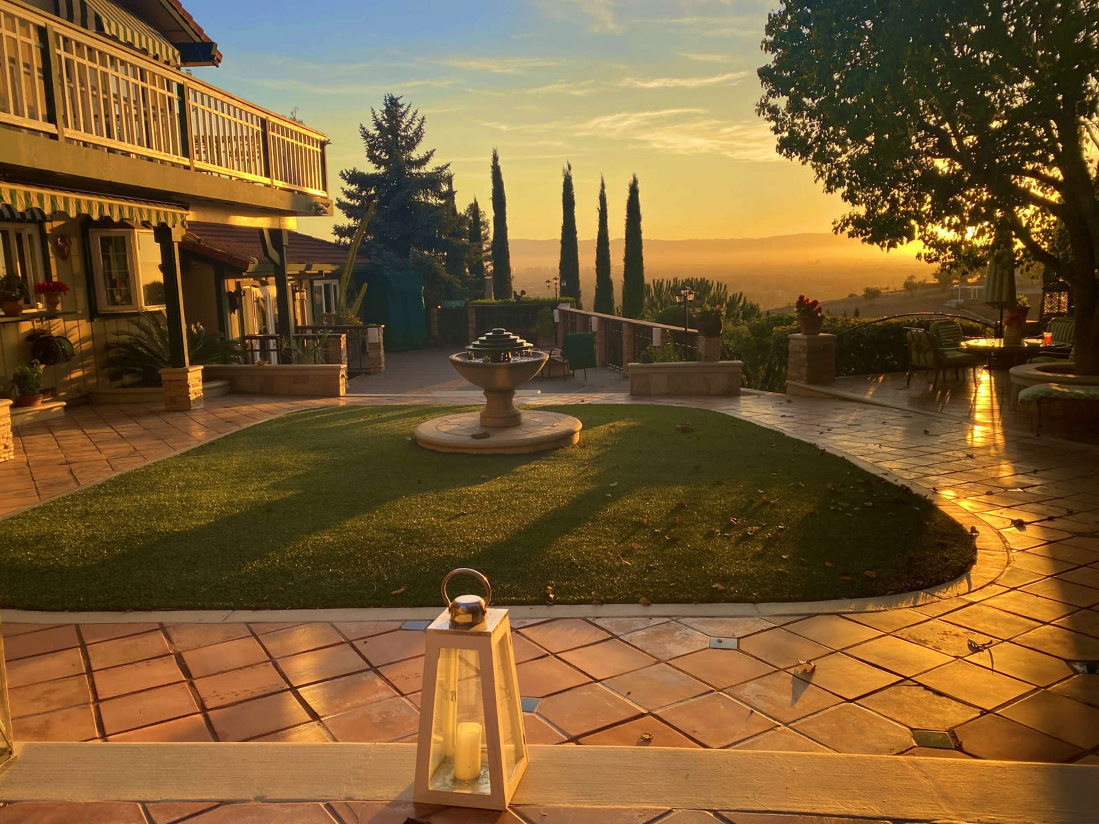 A decorative lantern sits in the foreground of a landscaped patio with a fountain and tall trees under a sunset sky.