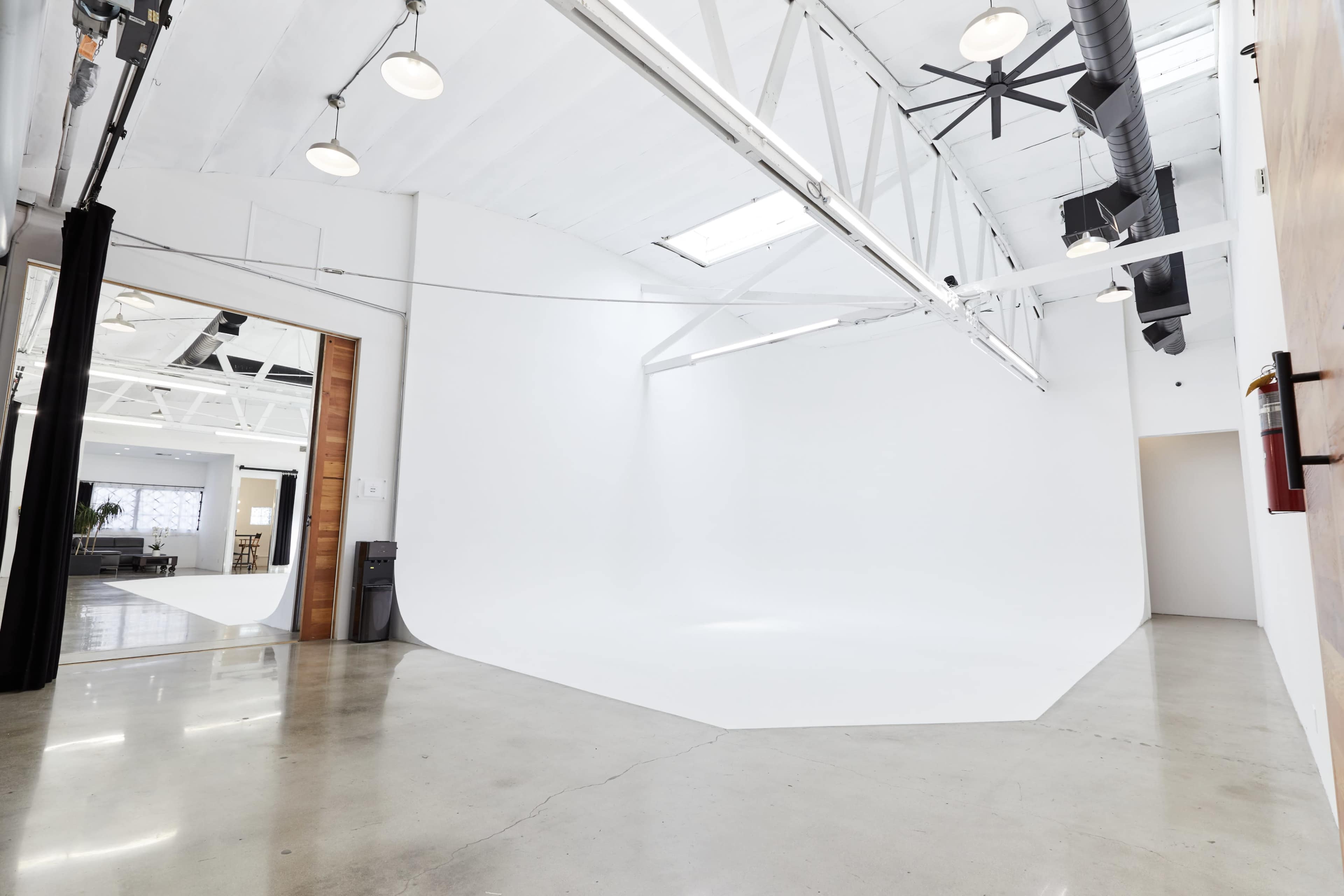 The image depicts a large, empty studio space with a seamless white backdrop, polished concrete floors, and industrial lighting.