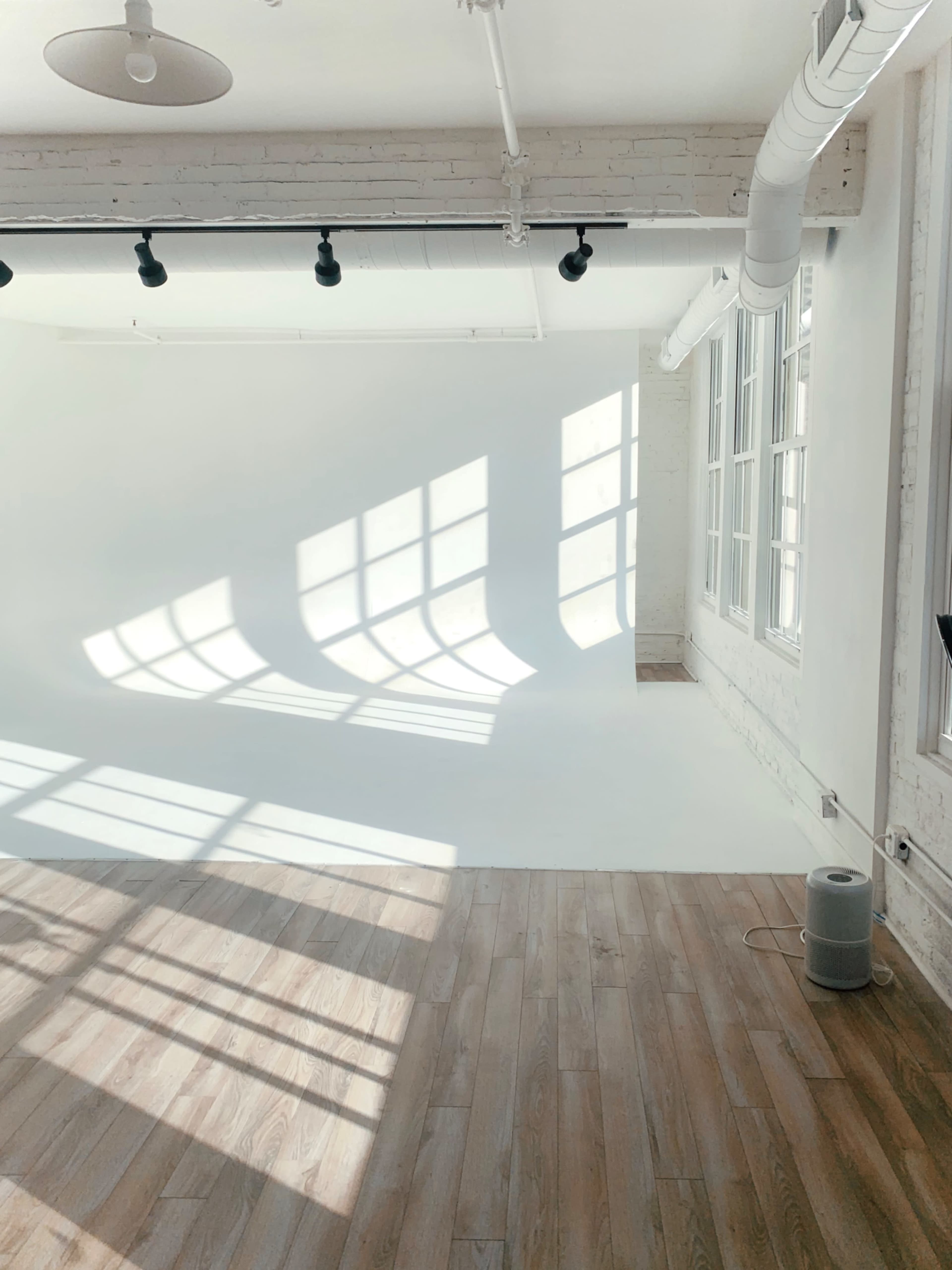 A bright, minimalist studio with large windows casting geometric shadows on a white backdrop and wooden floor.
