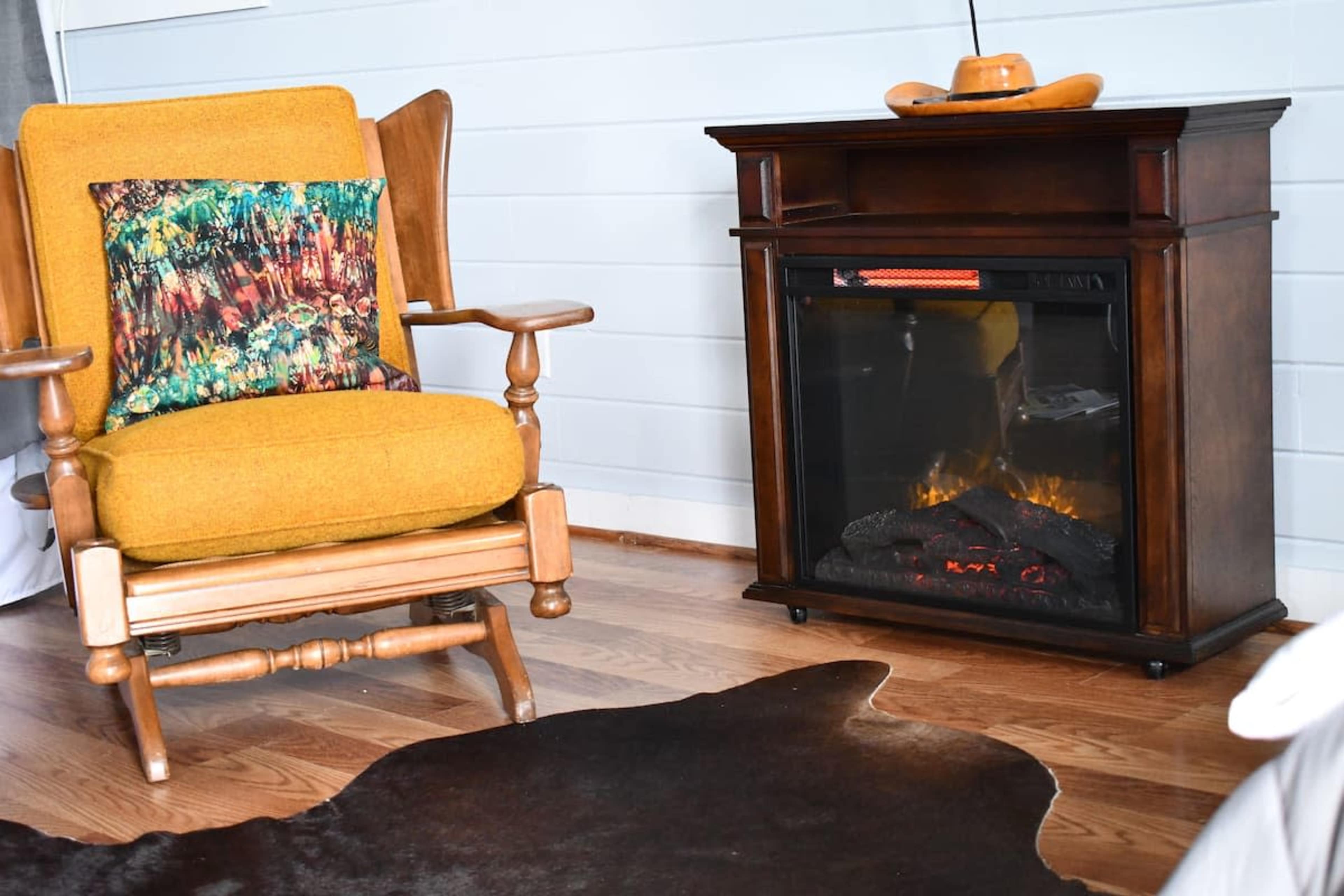 A rocking chair with a bright yellow cushion sits beside a wooden fireplace, with a cowhide rug on the floor.