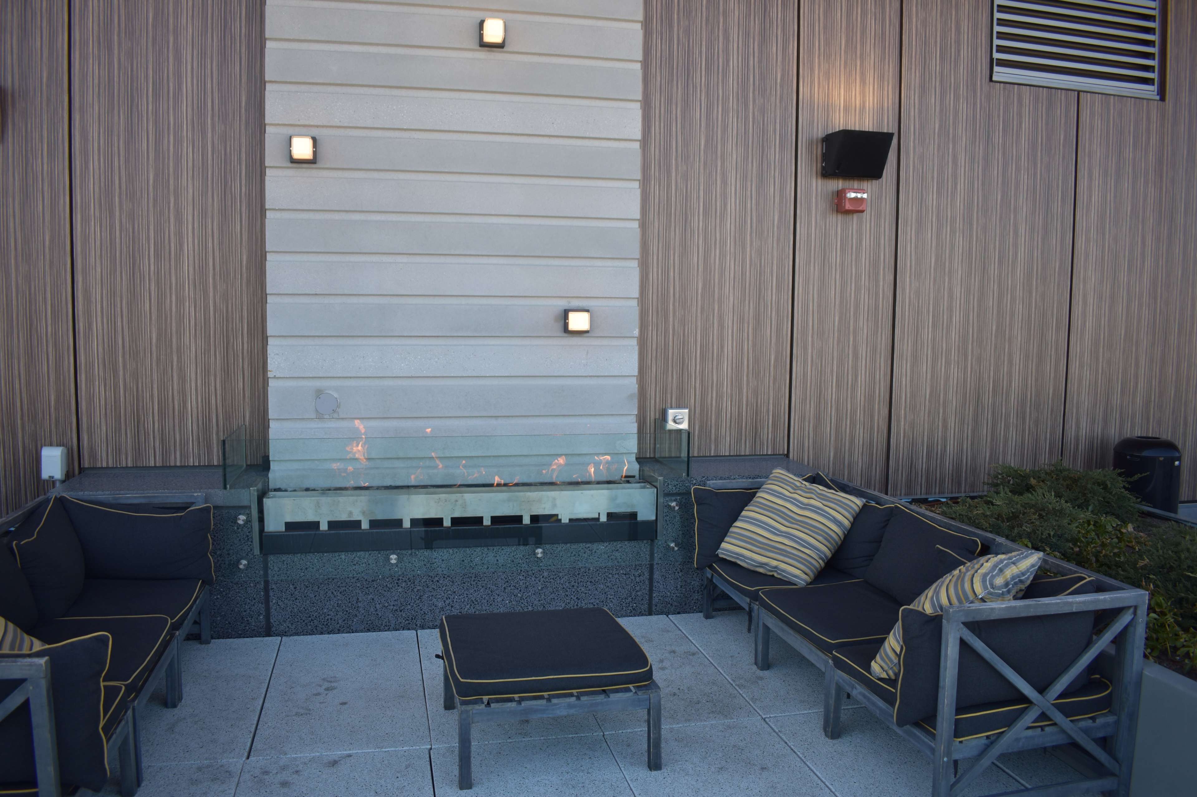 A seating area featuring gray outdoor furniture arranged around a linear fire feature against a wooden wall.