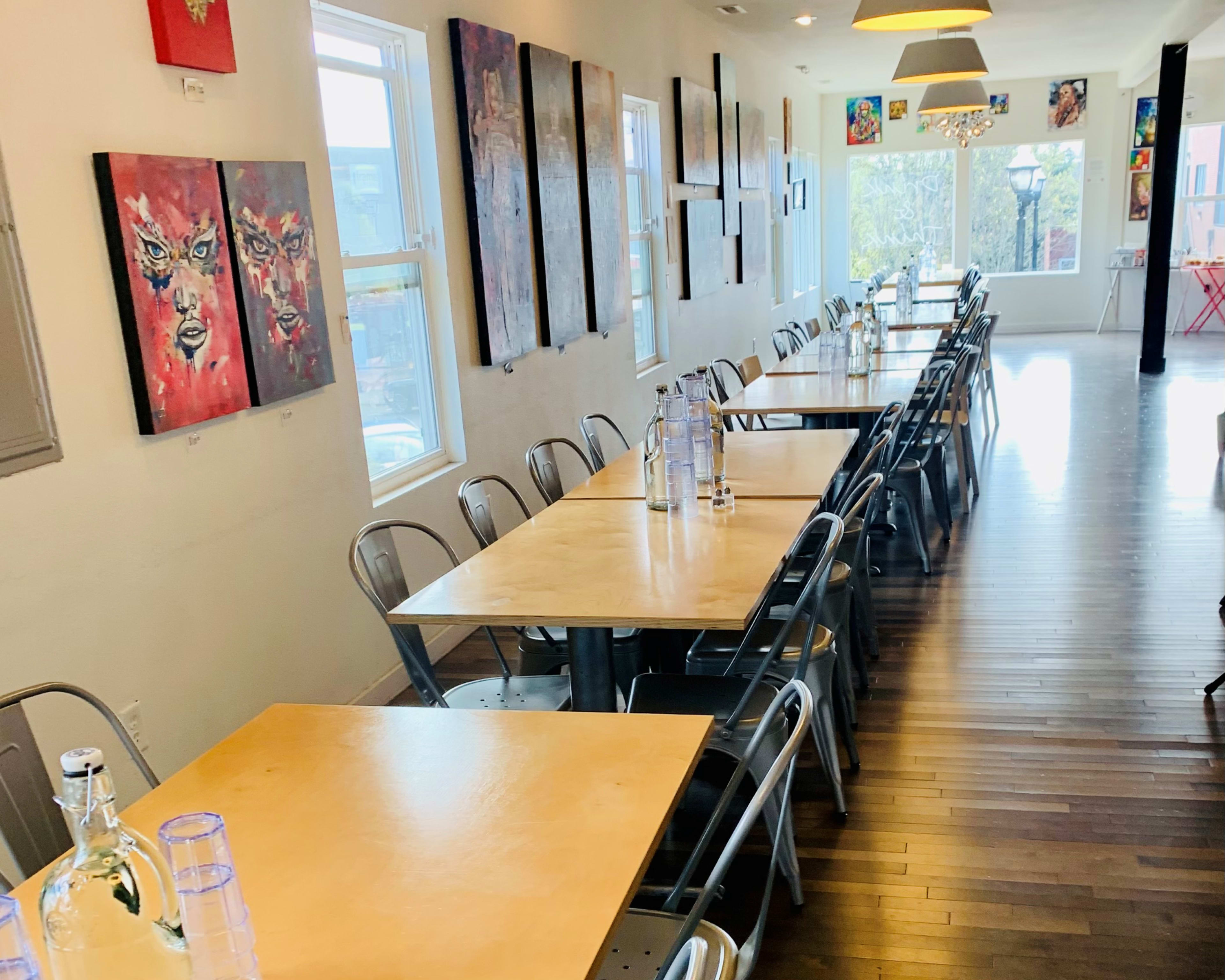 A long dining area with metal chairs and wooden tables, adorned with colorful artwork on the walls and large windows letting in natural light.