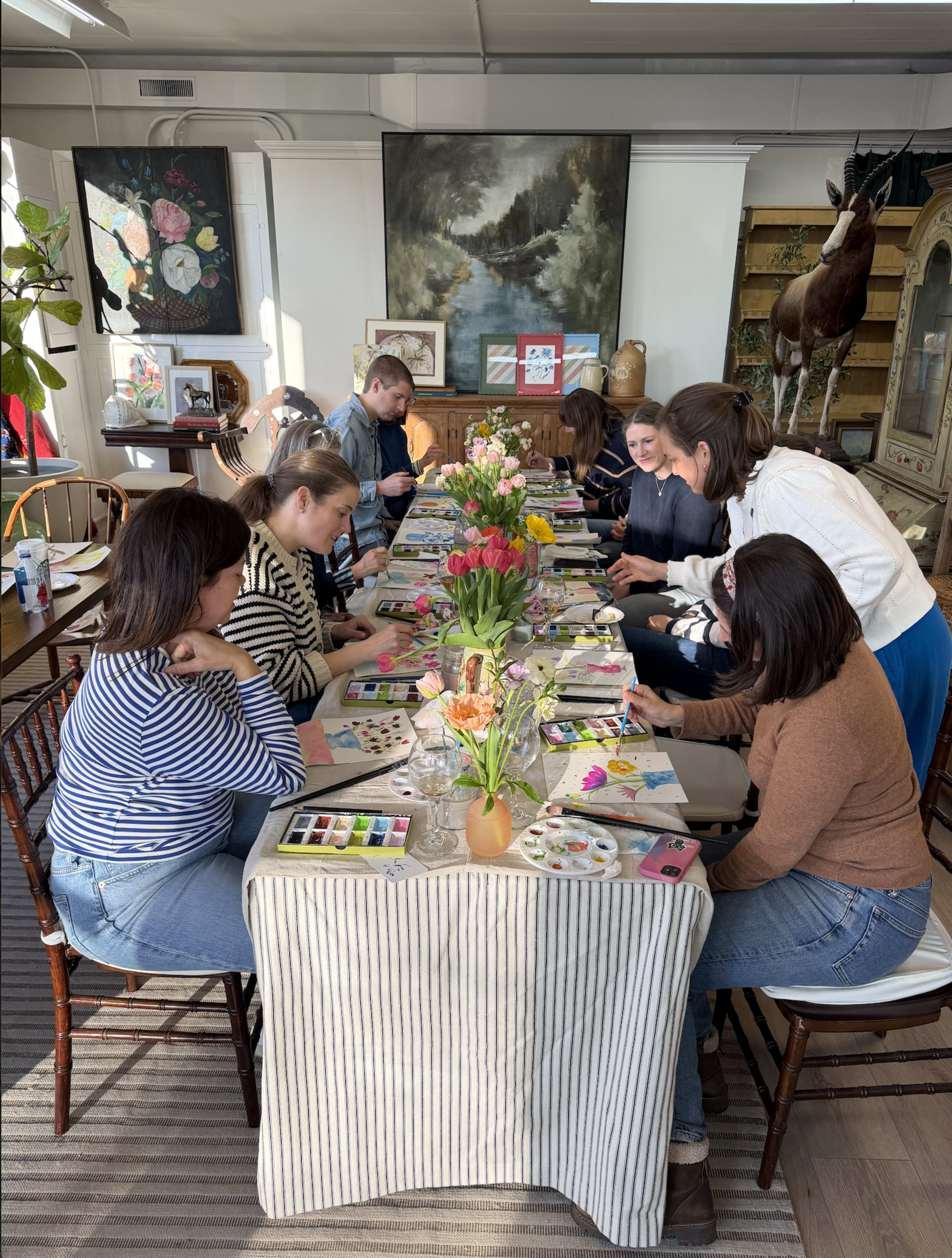 A group of individuals is seated around a long table adorned with art supplies, flowers, and colorful paintings in a well-lit room.
